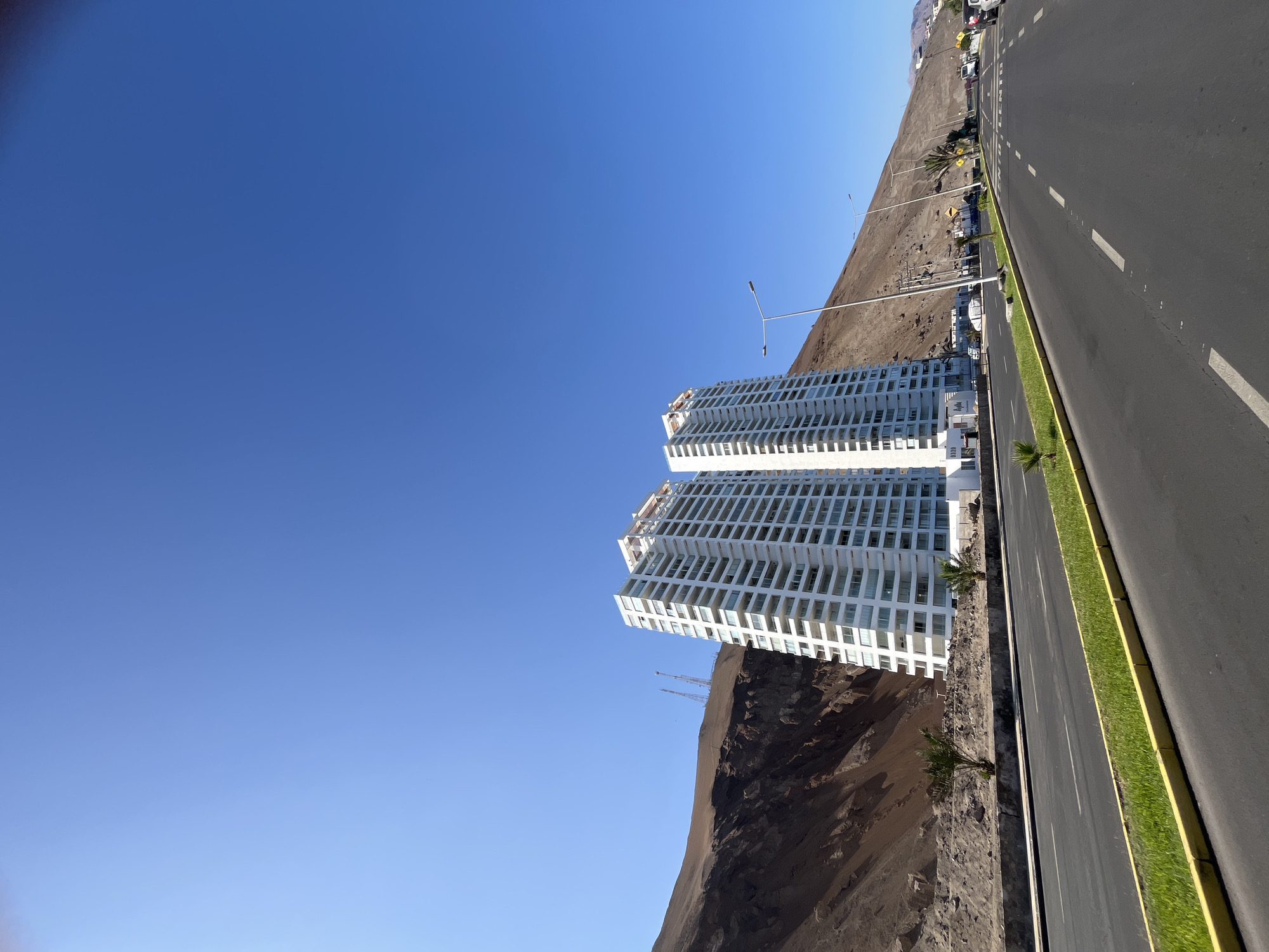 The image displays a clear, sunny daytime scene of a coastal urban landscape, likely in Arica, Chile, as provided by the context. The photograph is oriented vertically, showing a wide, multi-lane asphalt road extending diagonally from the foreground towards the upper right. The road features white dashed lane markings and a solid yellow line bordering a narrow, grassy median strip with small palm trees. Faint white text, possibly part of "BICENTENARIO," is visible on the road surface further down.
To the left of the road stand two prominent, tall, white residential buildings with a modern architectural design, characterized by many balconies and windows arranged in a grid-like pattern. These apartment towers are situated at the base of a steep, barren, reddish-brown hillside or mountain (cerro), which dominates the background to the left of the road. The hillside appears arid and rocky, with sparse vegetation. Some construction cranes are partially visible on the hillside behind the buildings.
The sky is a vibrant, cloudless blue, indicative of clear weather. Streetlights are positioned along the roadside, and a few small vehicles can be seen in the distance on the road. The scene captures a juxtaposition of modern urban development with a striking natural desert environment.