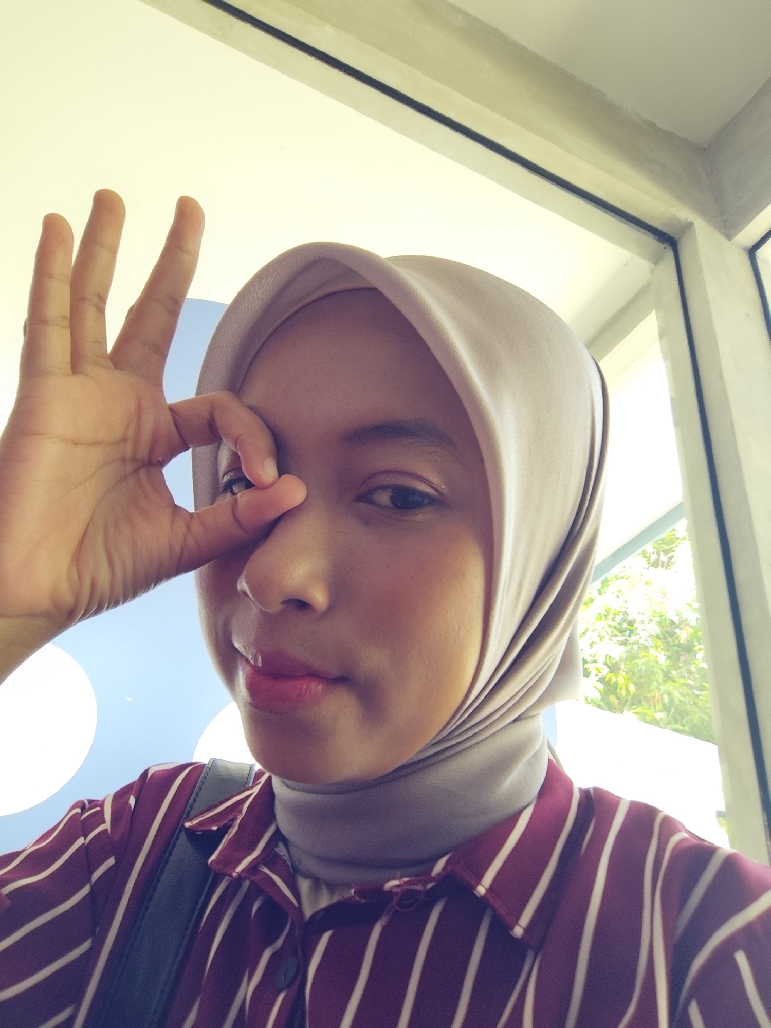 A close-up, waist-up shot shows a young woman wearing a beige hijab and a maroon and white striped collared shirt. She is looking at the camera and holding up her right hand to her eye, with her thumb and index finger forming a circle. Her left hand is partially visible at the top left of the frame. The background includes a bright sky, a window with trees visible outside, and what appears to be an indoor ceiling. The lighting suggests it is daytime.