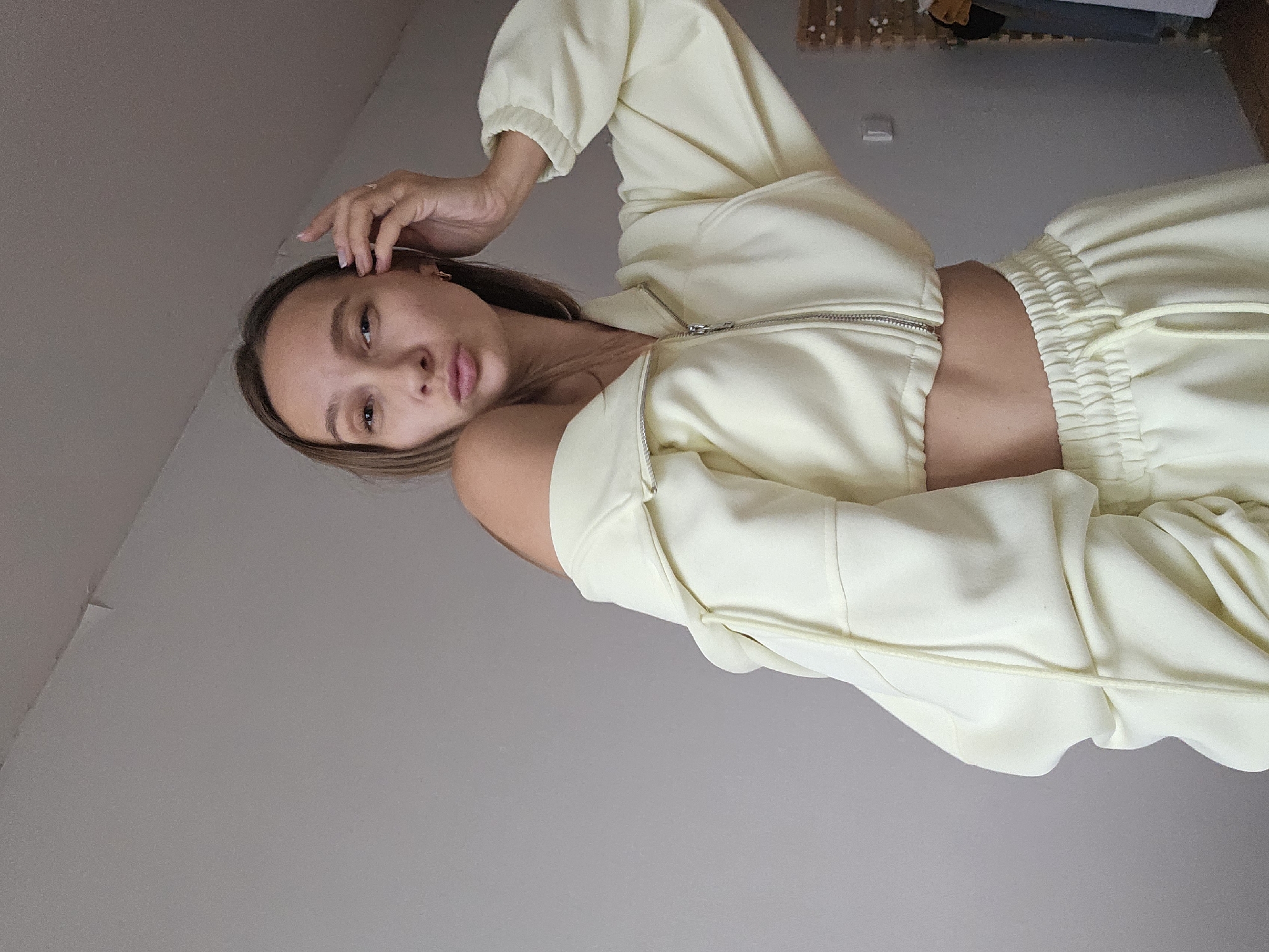 A young Caucasian woman with blonde hair and fair skin poses for a photo against a light gray wall. She is wearing a pale yellow cropped zip-up hoodie, with one shoulder exposed, and matching sweatpants. Her hand is raised to her head, and her lips are pursed. The background is sparsely furnished, with a hint of a closet in the upper right corner. The image is taken indoors, likely in a residential setting. There is no visible indication of the city of Chaikovsky, Russia.