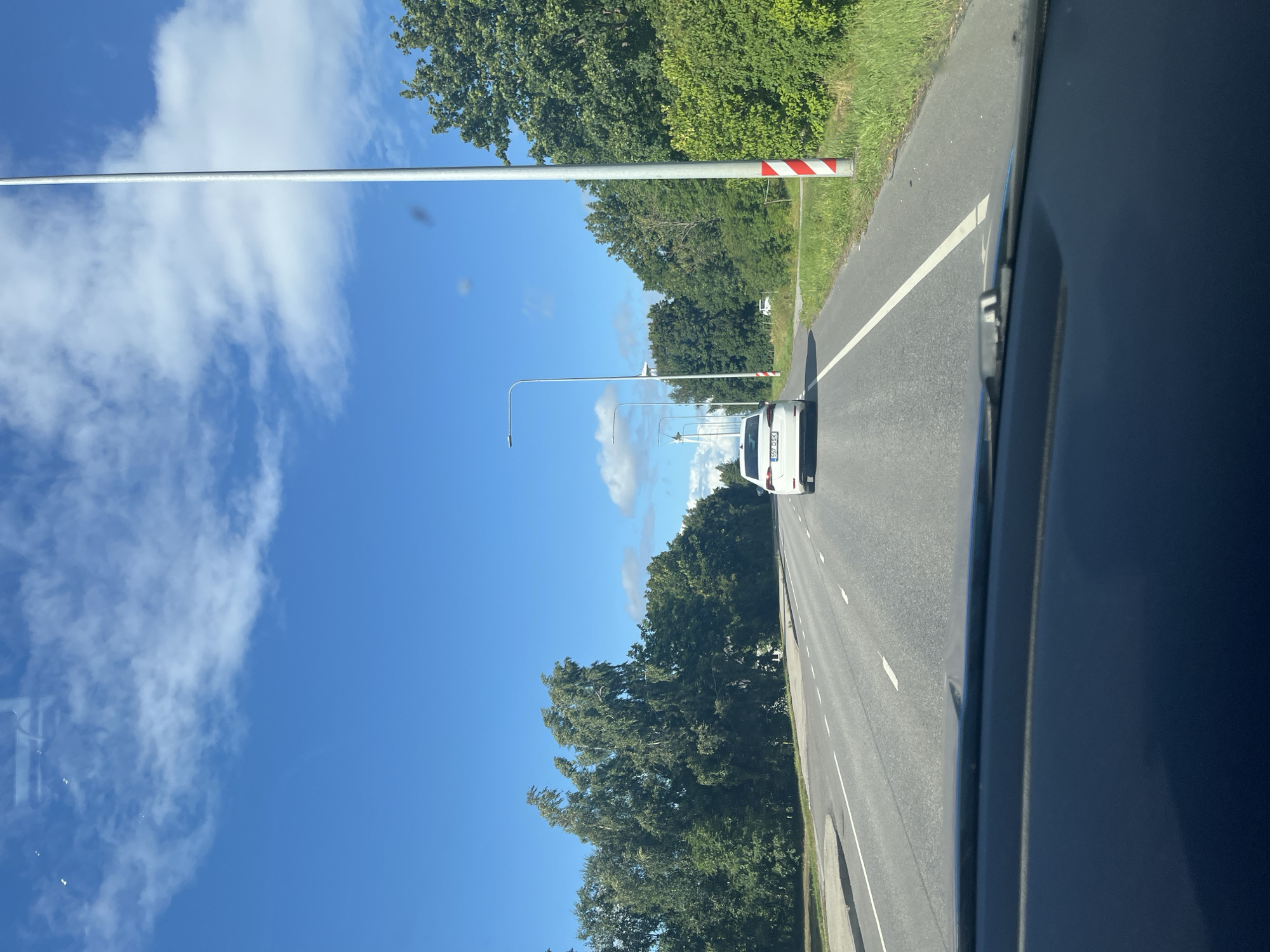 The image shows a view from inside a car looking forward on a two-lane road in Virtsu, Estonia. The sky is blue with scattered white clouds. Trees line both sides of the road, with lush green foliage. A white car with the license plate "557 DSX" is in front of the viewer's car, traveling in the same direction. There are streetlights visible along the road, and a tall pole with red and white markings stands on the right side. In the distance, a few white windmills can be seen. The road has white lane markings. The dashboard of the car is visible at the bottom of the image.