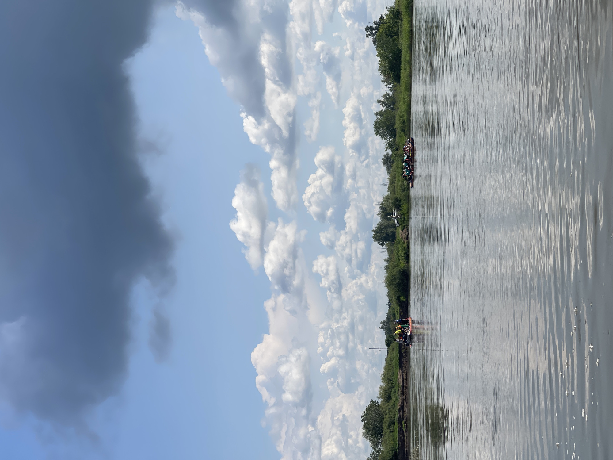 An aerial drone shot captures a wide, calm river flowing through a verdant landscape. The sky is a mosaic of bright blue and dramatic, dark grey clouds, suggesting an approaching storm. On the water, two rafts are visible. The closest raft, positioned in the middle of the frame, carries a group of people wearing life vests and holding paddles, actively engaged in kayaking. Further in the distance, a second raft, also populated by individuals in life vests, is further to the right. The banks of the river are lined with lush green trees and foliage. In the background, a few utility poles and the outline of a small aircraft can be discerned near the treeline, indicating proximity to a populated area or airfield.