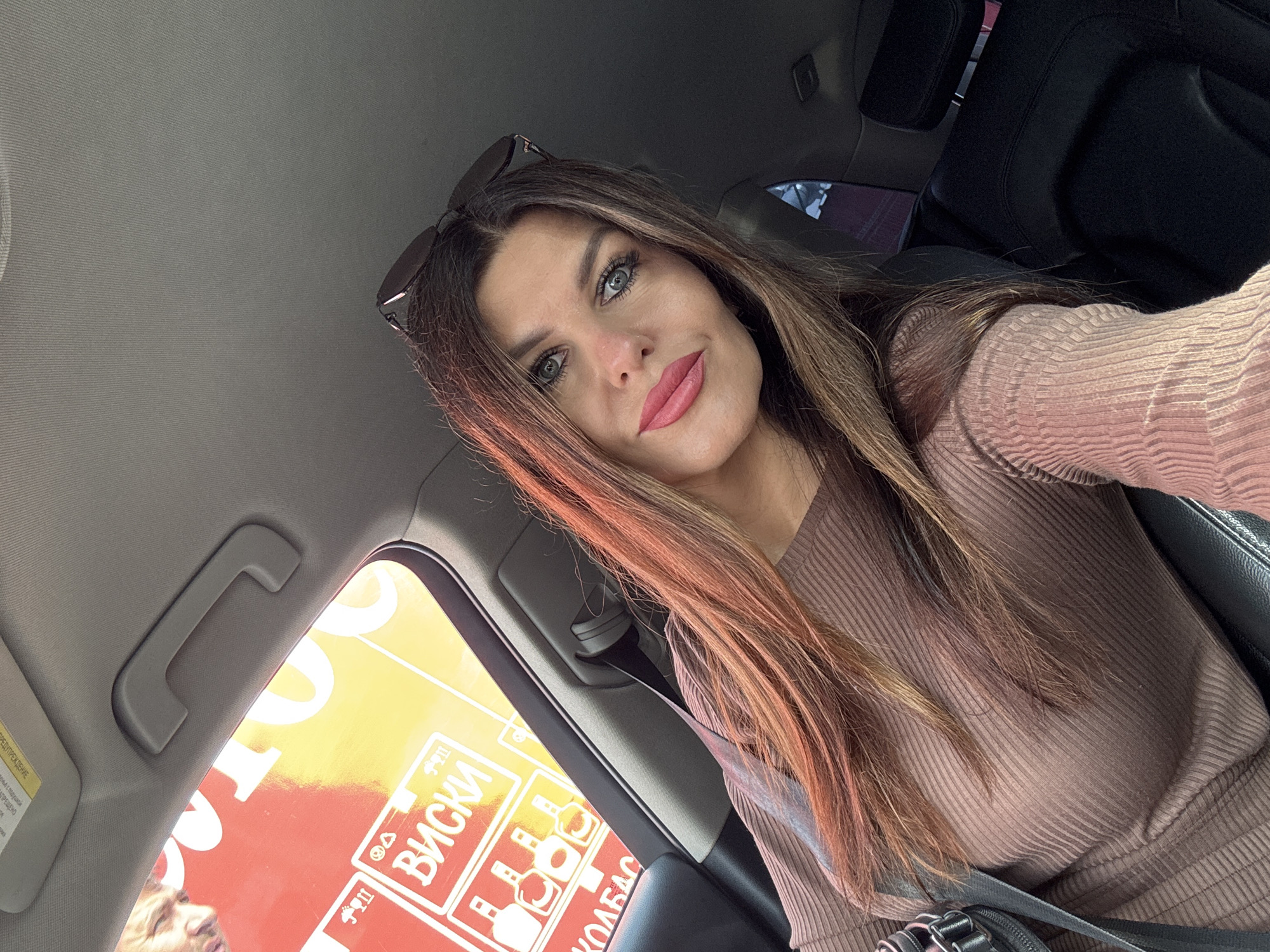 A woman is taking a selfie in a car. The woman has long brown hair with reddish highlights and is wearing a brown ribbed dress. She is wearing bright pink lipstick and has on sunglasses on top of her head. Through the car window, a sign with Russian text and images of bottles can be seen. The text on the sign says "ВИСКИ" (whiskey) and "КОЛБАС" (sausage). A man's face is partially visible in the upper left corner of the image, looking out the window. The interior of the car, including the ceiling handle and car seat, is also visible. The image appears to have been taken during the daytime.