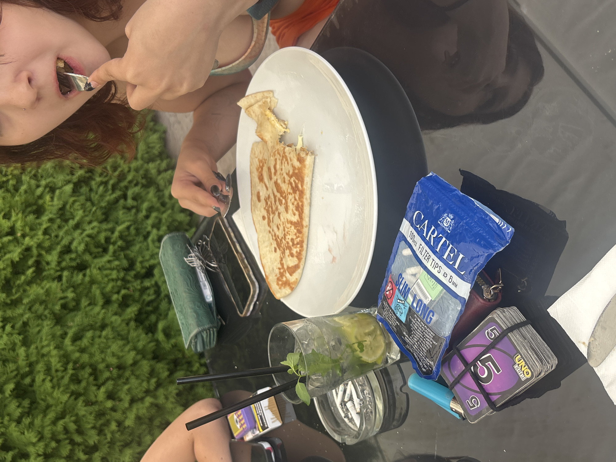 A medium shot captures a table set for a meal outdoors, with lush green foliage forming a backdrop. A person, only their upper body visible, is in the process of eating from a plate of food with a fork. On the table, a drink with mint and lime sits in a glass, alongside an ashtray filled with cigarette butts, a pack of cigarettes, a blue lighter, a stack of UNO cards, a wallet, and a package of filter tips. A smartphone lies flat on the table, partially obscured by a green, textured object. The scene is set on a dark, reflective surface, suggesting a table outdoors. The image does not provide enough information to determine the specific location within Zverino, Bulgaria.