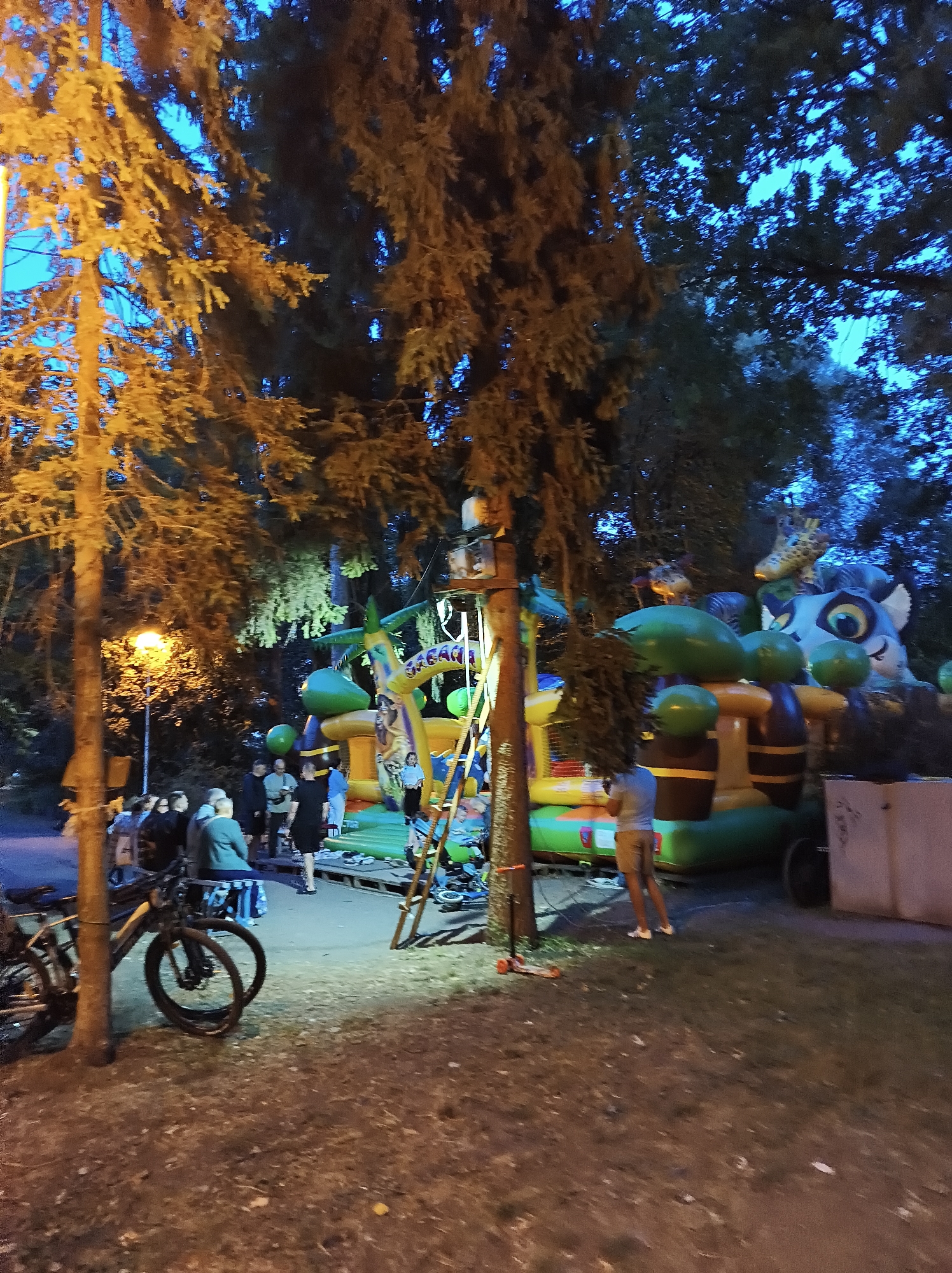 An outdoor scene at dusk or night in Ternopil, Ukraine, features a lively gathering around a large inflatable attraction with a jungle theme. The inflatable structure, predominantly green and yellow, is illuminated and depicts cartoon animals like giraffes and a large cat's face. Several people are visible, some standing and conversing, while others appear to be children or adults interacting with the attraction or observing. A bicycle is parked near a tree in the foreground, and a lamppost emits a warm glow, illuminating the area. The background is filled with trees, creating a natural setting. The ground is covered with grass and scattered leaves, suggesting a park or an open outdoor space. The overall atmosphere is one of community enjoyment and nighttime recreation.