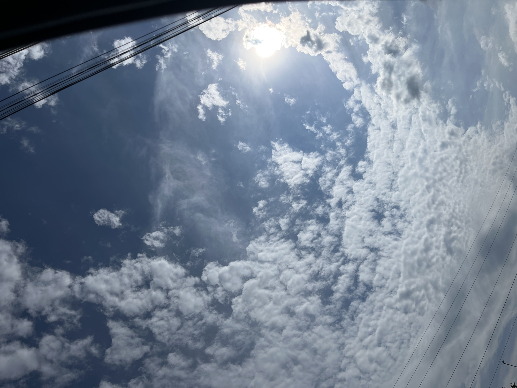 The image shows a view of the sky with scattered clouds. The sun is visible in the upper right quadrant, partially obscured by clouds, casting a bright glare. Several thin, dark power lines stretch diagonally across the frame from the bottom left towards the upper right. The scene appears to be outdoors, possibly in a semi-urban or rural area given the presence of power lines. There are no people or discernable structures in the immediate foreground or background. The sky is a deep blue, indicating daylight.