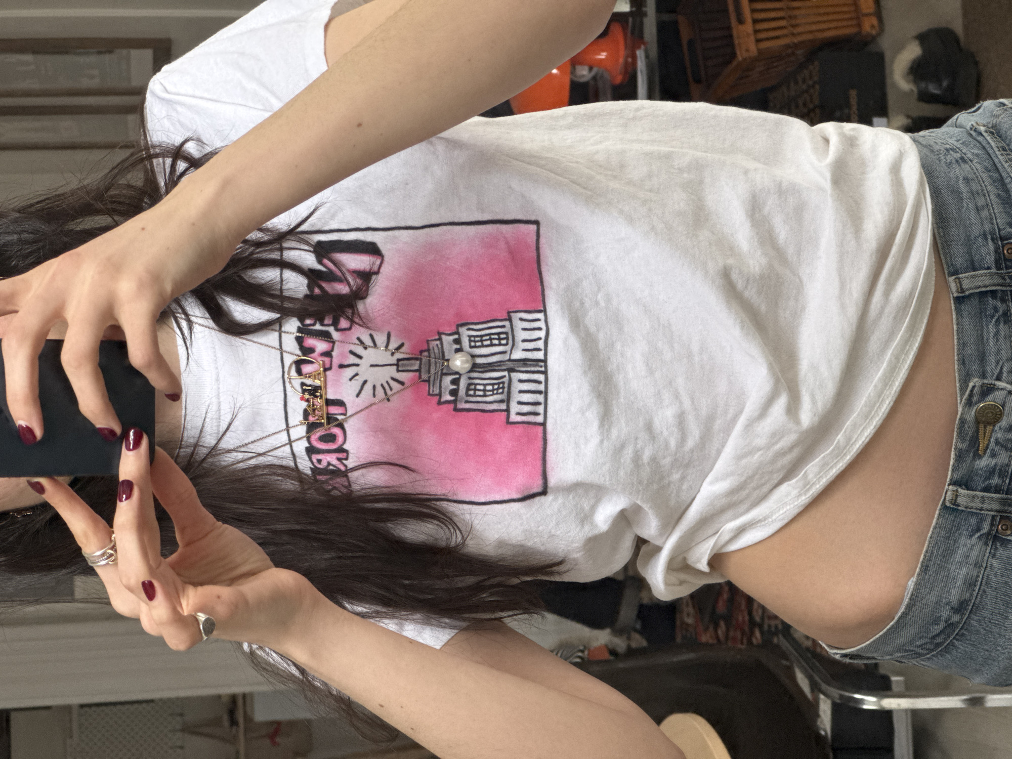 A woman with long dark hair is holding a phone in her hands, taking a selfie. She is wearing a white t-shirt with a graphic design on it that includes an image of the Empire State Building. She is also wearing denim shorts. The location is not discernible.