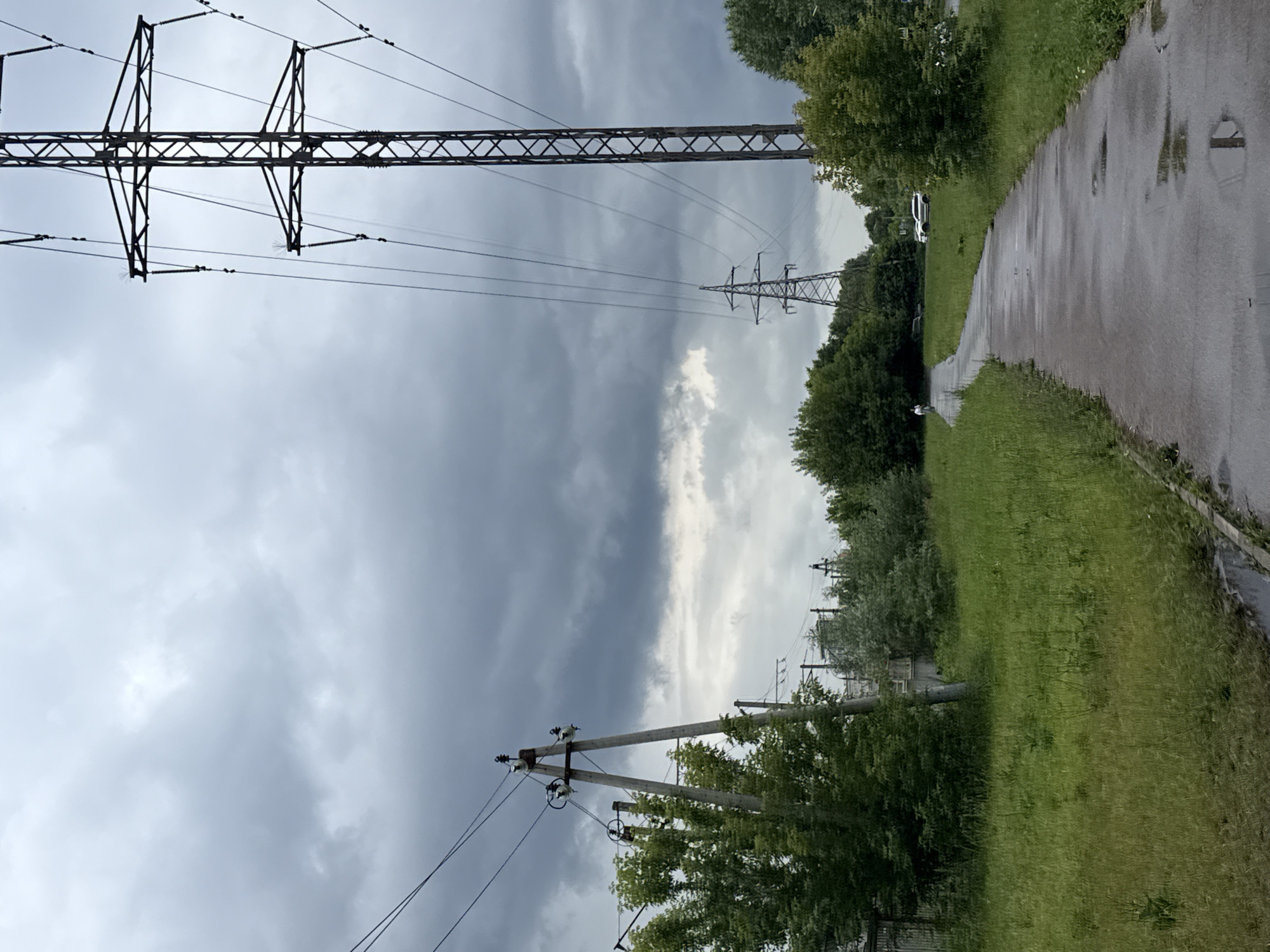 This media file displays a scene with electrical infrastructure against a cloudy sky. Dominating the left side of the frame are two large metal power pylons and a smaller wooden utility pole. The pylons are tall, lattice structures with numerous wires strung between them. The utility pole is closer to the foreground, featuring insulators and wires.
To the right, a wet asphalt pathway runs alongside a grassy area. The pathway shows reflections, suggesting recent rain. Trees and lush green grass line the path and surround the electrical infrastructure. In the distance, a white car is visible on the pathway. A person is walking on the pathway in the middle ground.
The sky is filled with heavy, gray clouds, indicating overcast weather, possibly before or after a rain shower. The lighting is dim and diffused. The overall mood is somewhat somber due to the cloudy conditions and the presence of industrial infrastructure. There is no visible text in the image. The setting appears to be an outdoor, possibly semi-rural or suburban area with established infrastructure. The time of day is not precisely indicated but the lighting suggests it is daytime.