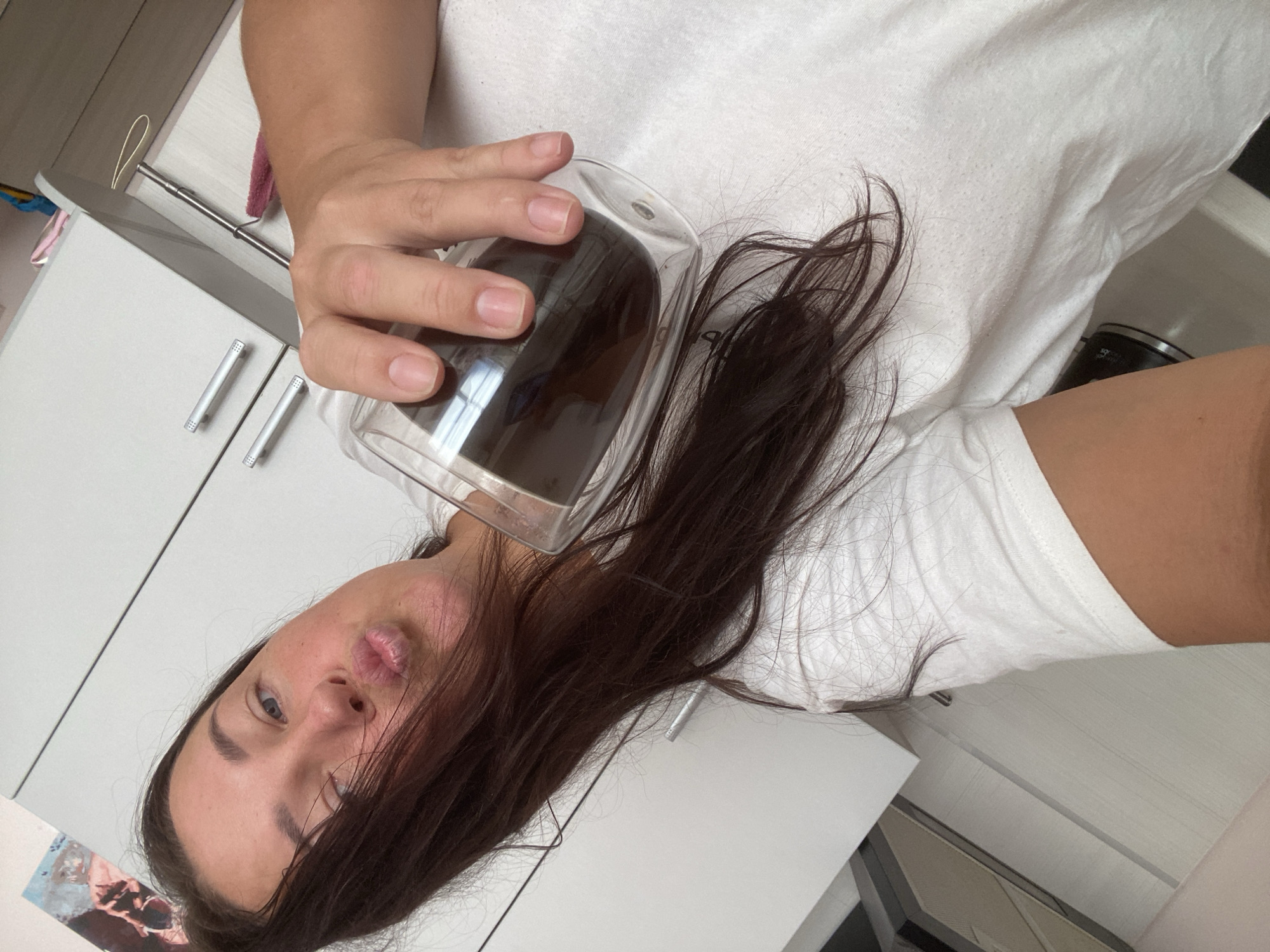 A woman is taking a selfie in a kitchen. She is wearing a white t-shirt and has her hair down. She is holding a glass of dark liquid in her right hand and puckering her lips towards the camera. The kitchen has white cabinets and stainless steel handles. The image is taken from a low angle, looking up at the woman. There is no visible indication of the location being in Sochi, Russia.