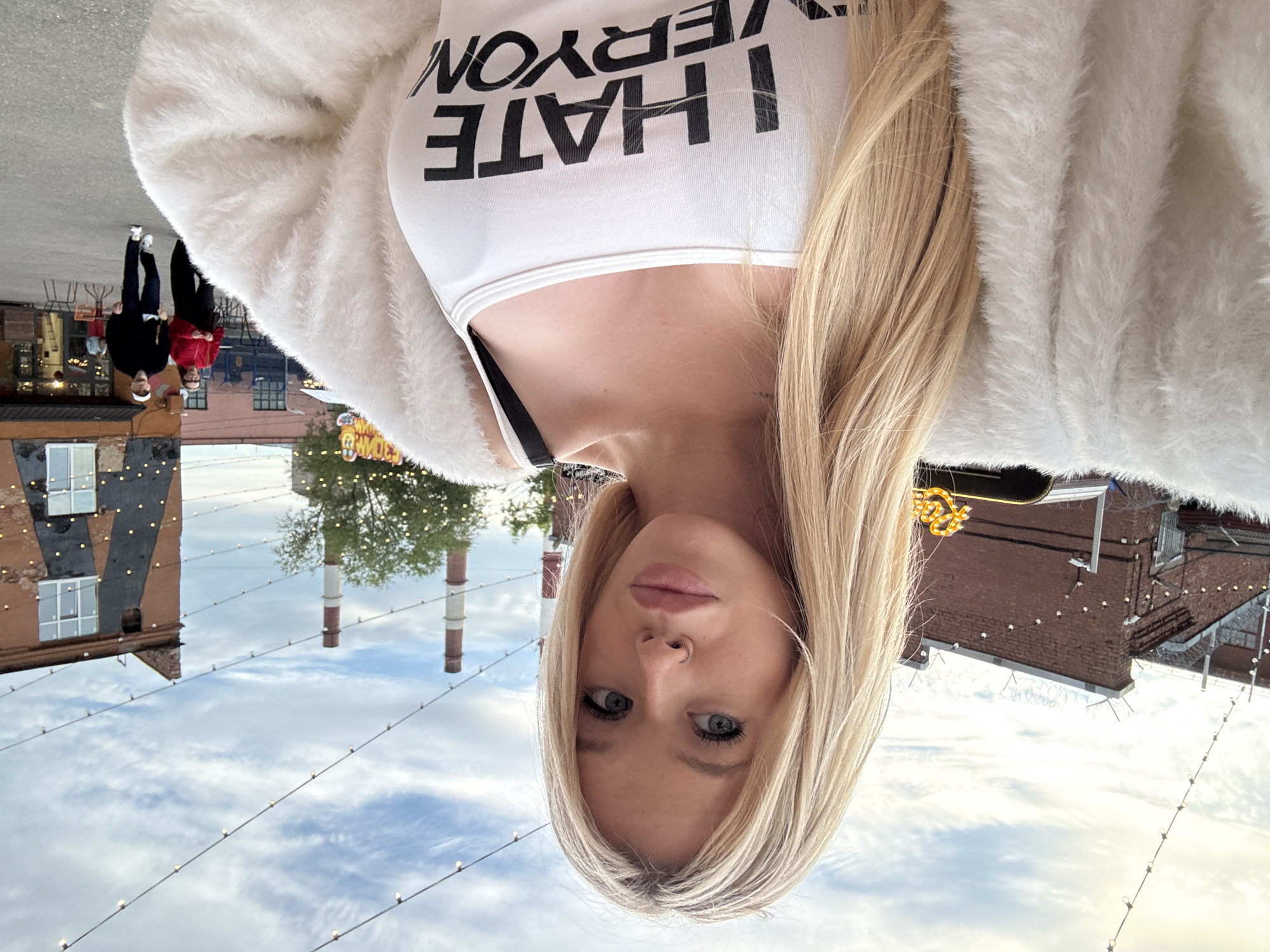The image is a selfie taken outdoors in Saint Petersburg, Russia. In the foreground, a young Caucasian woman with blonde hair and blue eyes is visible from the chest up. She is wearing a white tank top with the black text "I HATE EVERYON" printed on it, and a fluffy white jacket. The background shows a street scene with brick buildings, industrial chimneys, trees, and string lights overhead. Two people, a man and a woman, are walking away from the camera in the middle ground. The overall setting appears to be a public area, possibly a market or entertainment district. The sky is partly cloudy.