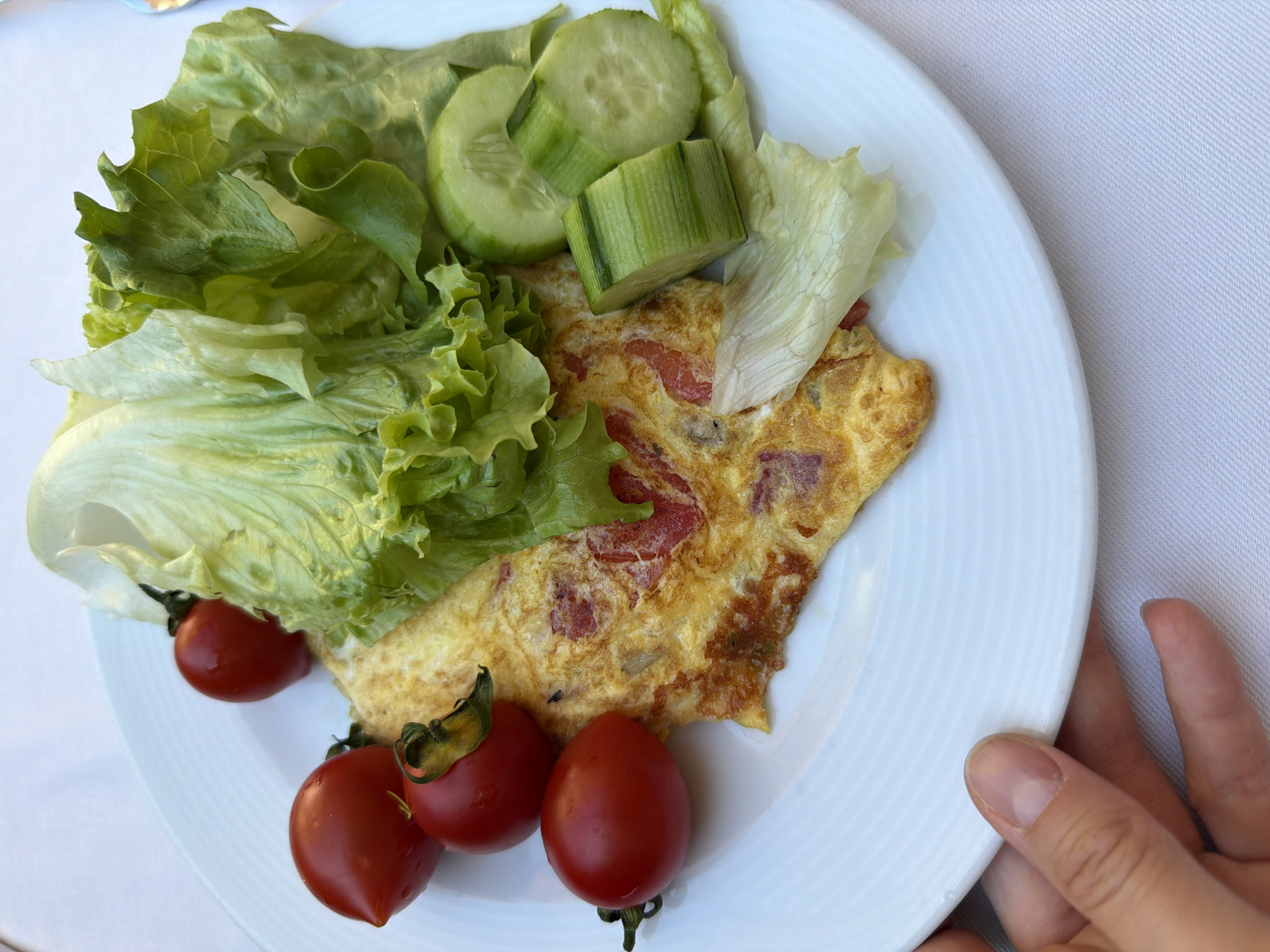 A top-down shot shows a white plate holding a breakfast meal consisting of a tomato omelet, a pile of green lettuce, sliced cucumbers, and three cherry tomatoes with stems. The omelet has brown edges and red tomato chunks embedded within. The cucumber slices are circular and rectangular. The lettuce is a mix of dark and light green leaves. The plate has concentric ridges along its rim. A person's hand with neatly trimmed nails is visible on the right side of the image, holding the edge of the plate. The background appears to be a white tablecloth. This breakfast meal is likely being served in a location in Fethiye, Turkey.