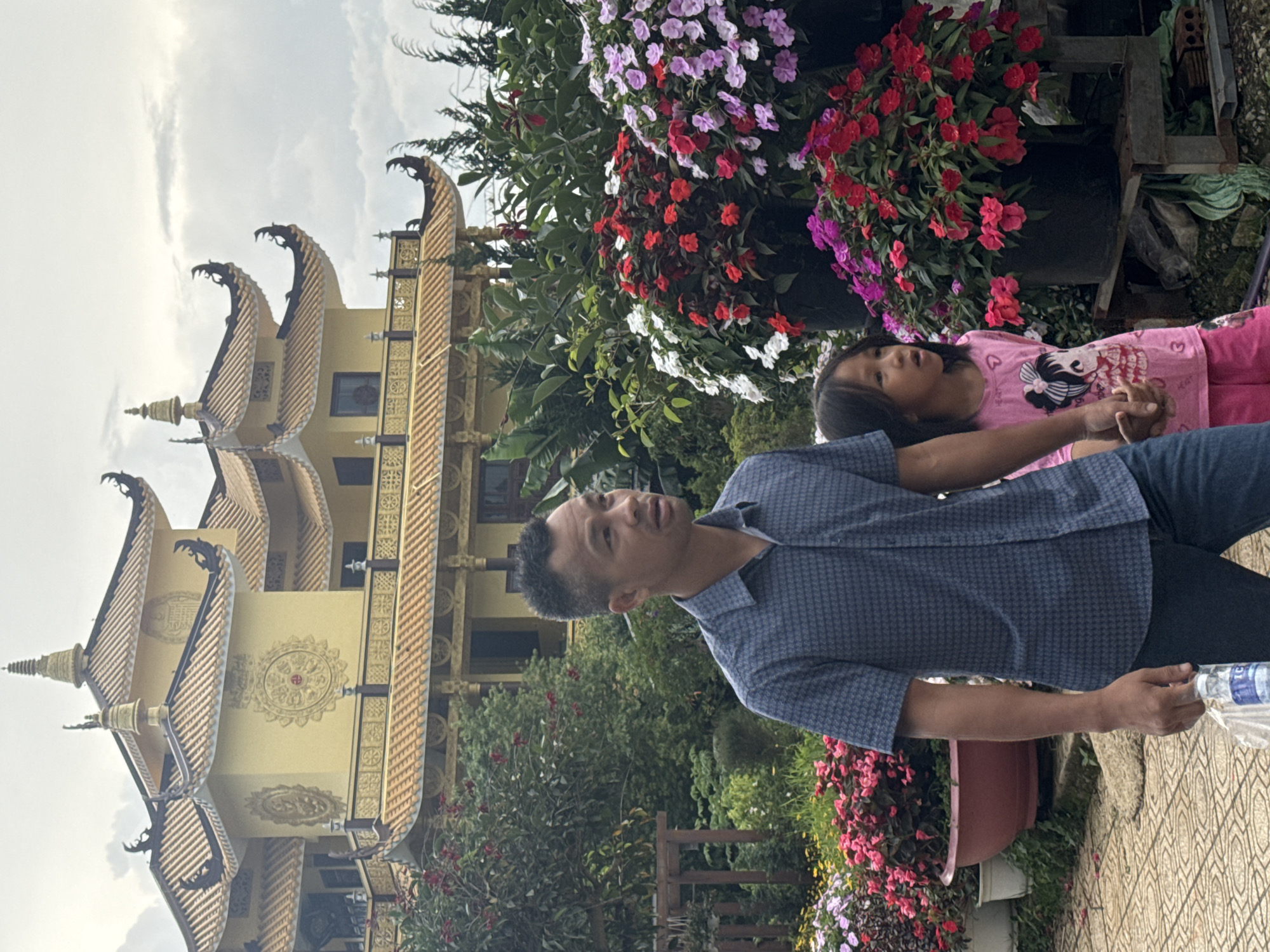 The image depicts a father and daughter walking past a vibrant display of flowers in front of a yellow Buddhist temple. The temple, with its intricate golden roof details and circular decorative motifs, is characteristic of traditional Vietnamese temple architecture and suggests a location within or near a religious site in Da Lat, Vietnam. The father, wearing a blue patterned short-sleeved shirt and jeans, is on the left, holding a plastic bag containing a water bottle. The daughter, on the right, wears a pink t-shirt with a graphic print and pink shorts, and is holding her father's hand. They are walking on a tiled pathway surrounded by lush greenery and numerous pots overflowing with colorful flowers, including red, pink, and white impatiens. The background is filled with dense foliage and the imposing structure of the temple, under a cloudy sky. The overall scene conveys a sense of familial outing in a culturally rich and visually appealing environment.
