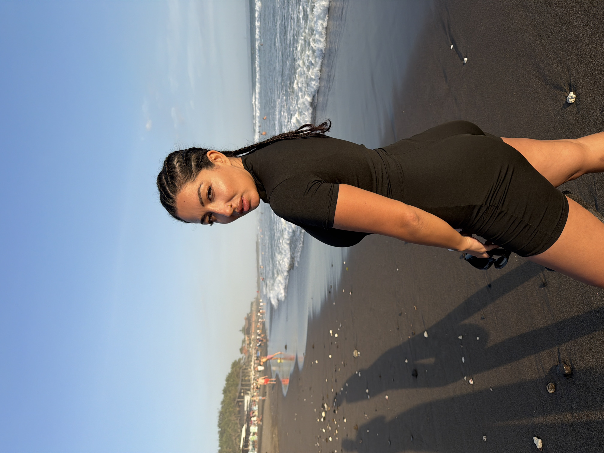 A woman with braided hair stands on a black sand beach in North Kuta, Indonesia. She is wearing a black, form-fitting outfit and is looking over her shoulder at the camera. The ocean with gentle waves is to her right, and a line of trees and buildings is visible in the distance along the coastline. Several other people are scattered along the beach and in the water. The sky is a clear blue with a soft gradient, indicating it is likely late afternoon. Shadows of the woman and other people are cast on the dark sand.