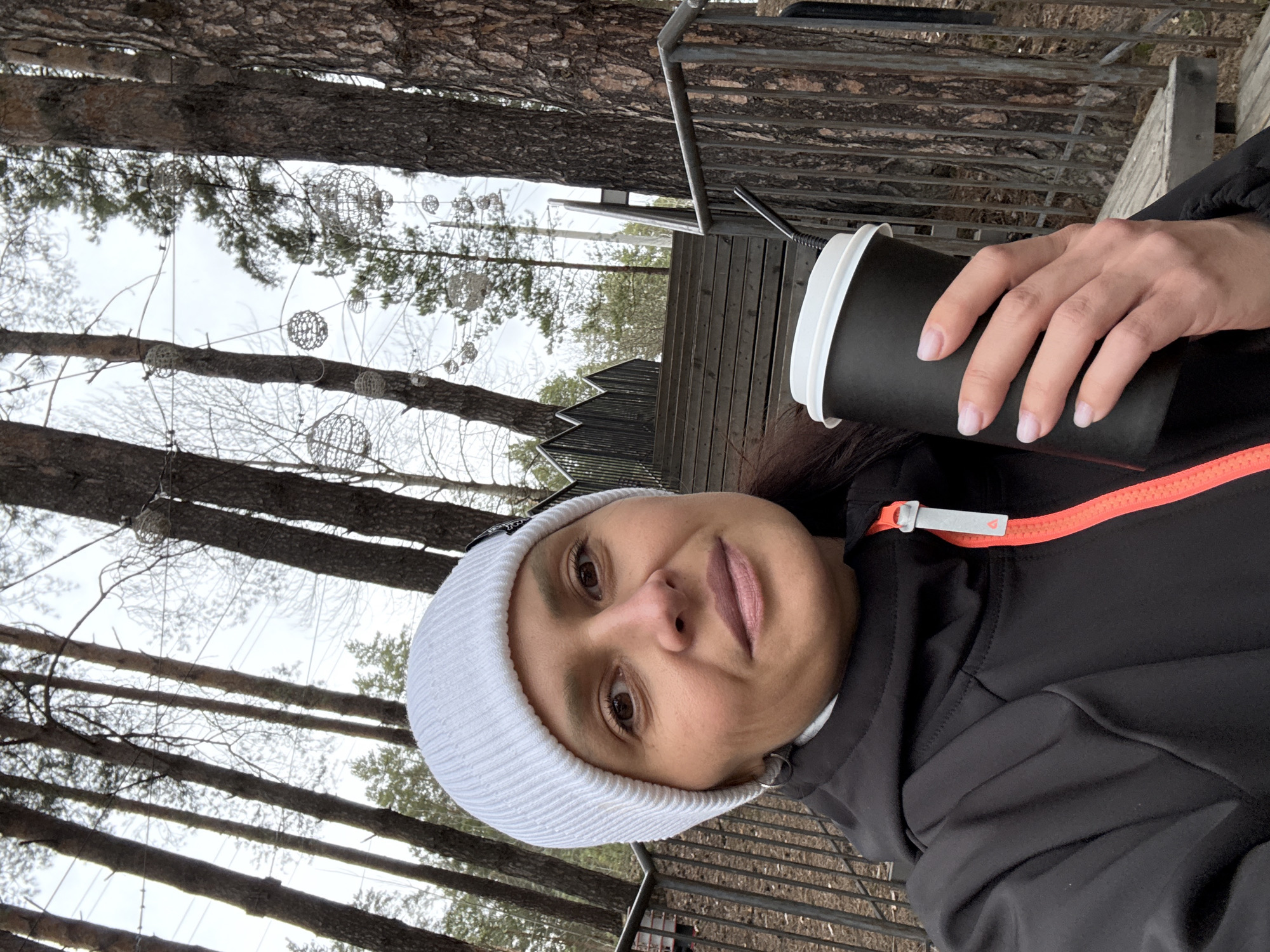 A woman wearing a white beanie and a black jacket with orange trim is holding a black disposable coffee cup with a white lid. The photo is taken outdoors in what appears to be a wooded area. Tall trees with brown bark surround her. Decorative wire orbs are strung between the trees. A metal railing and wooden benches are visible in the background. The image was taken in Neftekamsk, Russia.