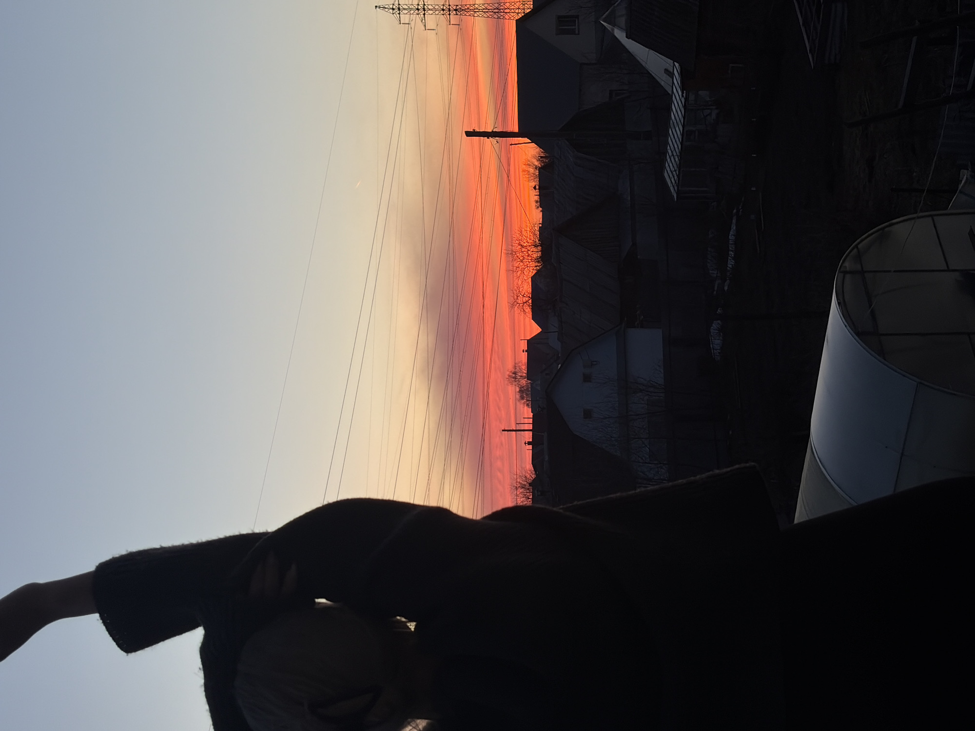 The image is a landscape photo taken in Chaikovsky, Russia, at sunset. The sky is multicolored, transitioning from blue to orange to red as it gets closer to the horizon. A power line and an electrical tower are visible against the sky. Below the horizon, there are silhouettes of houses and trees. In the foreground, a person is partially visible, obscured in shadow, with only their shoulder and head discernible. A greenhouse is situated to the right side of the view. The lighting suggests it is either dawn or dusk, and the overall mood of the scene is tranquil.