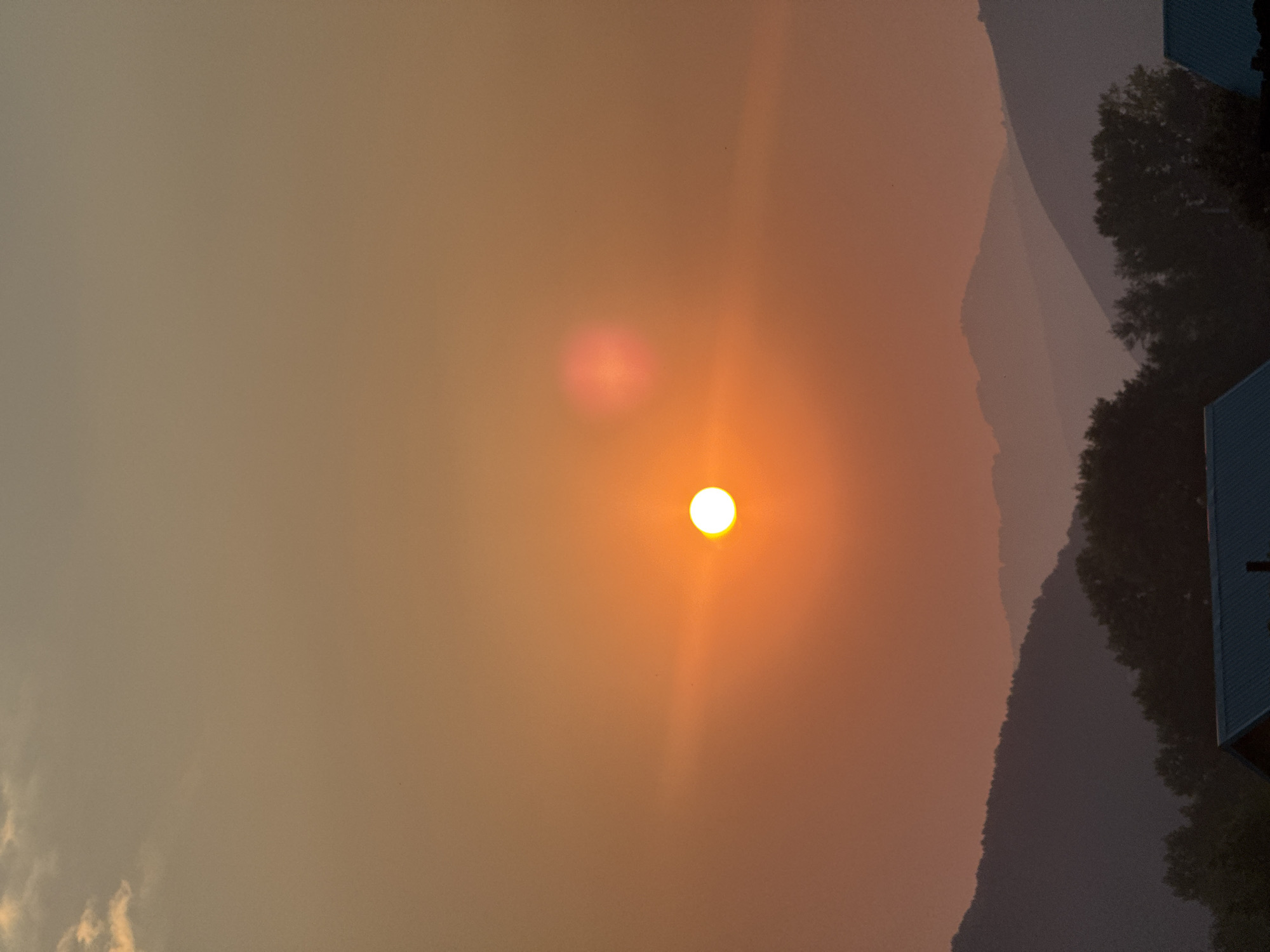 A vertical, medium shot captures a sunset over a landscape, likely in the city of Soloneshnoye, Russia. The sun, a bright, incandescent orb of orange, is positioned slightly left of the vertical center, casting a warm, diffused glow across the hazy sky. A fainter, reddish lens flare is visible above and to the right of the sun. In the foreground, silhouetted against the sunset, are layered, dark mountain ranges, their peaks softened by distance and atmosphere. Below the mountains, the dark silhouettes of trees form a dense band. At the very bottom of the frame, the dark roofs of several buildings, possibly houses, are visible. The overall impression is one of a serene, natural landscape bathed in the dramatic light of twilight.