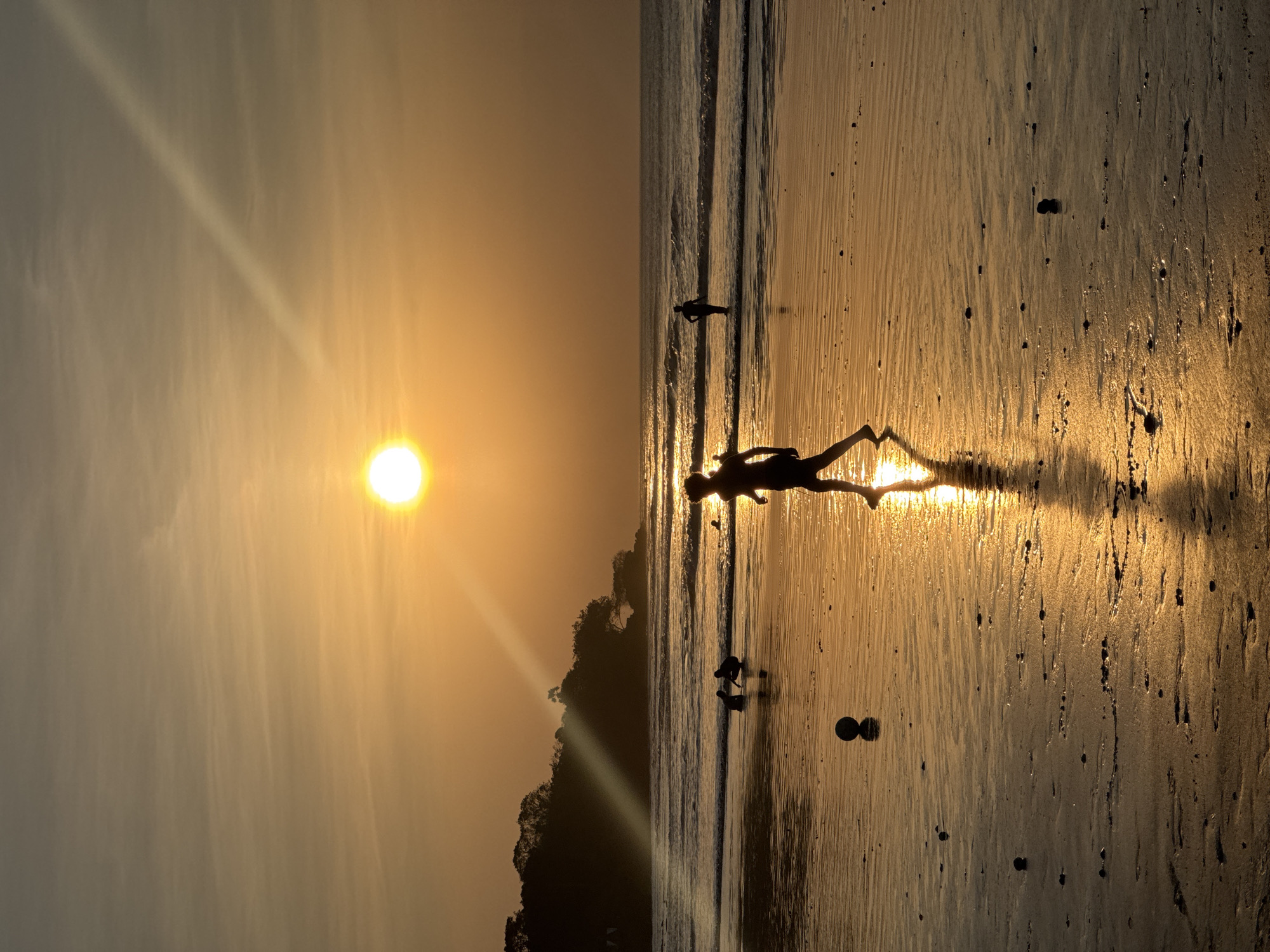 The image depicts a serene sunset scene on Kuta Selatan beach, Indonesia. The sun, a bright orb of golden light, hangs low in the sky, casting a warm, orange glow across the horizon and the wet sand. The sun's rays create a luminous path across the water and the beach, highlighting the textures of the wet sand. Several figures are present, silhouetted against the bright light. In the foreground, a lone individual walks across the sand, their reflection shimmering on the wet surface. To the left, two other figures are crouched near the water's edge, appearing to interact with the sand or a small ball. Further out in the ocean, another solitary figure stands in the shallow waves, their silhouette distinct against the backdrop of the sea and sky. The ocean is calm, with gentle waves rolling towards the shore. A dark, tree-covered headland is visible in the distance on the left side of the image, adding depth to the landscape. The overall atmosphere is peaceful and tranquil, capturing the beauty of a tropical sunset.