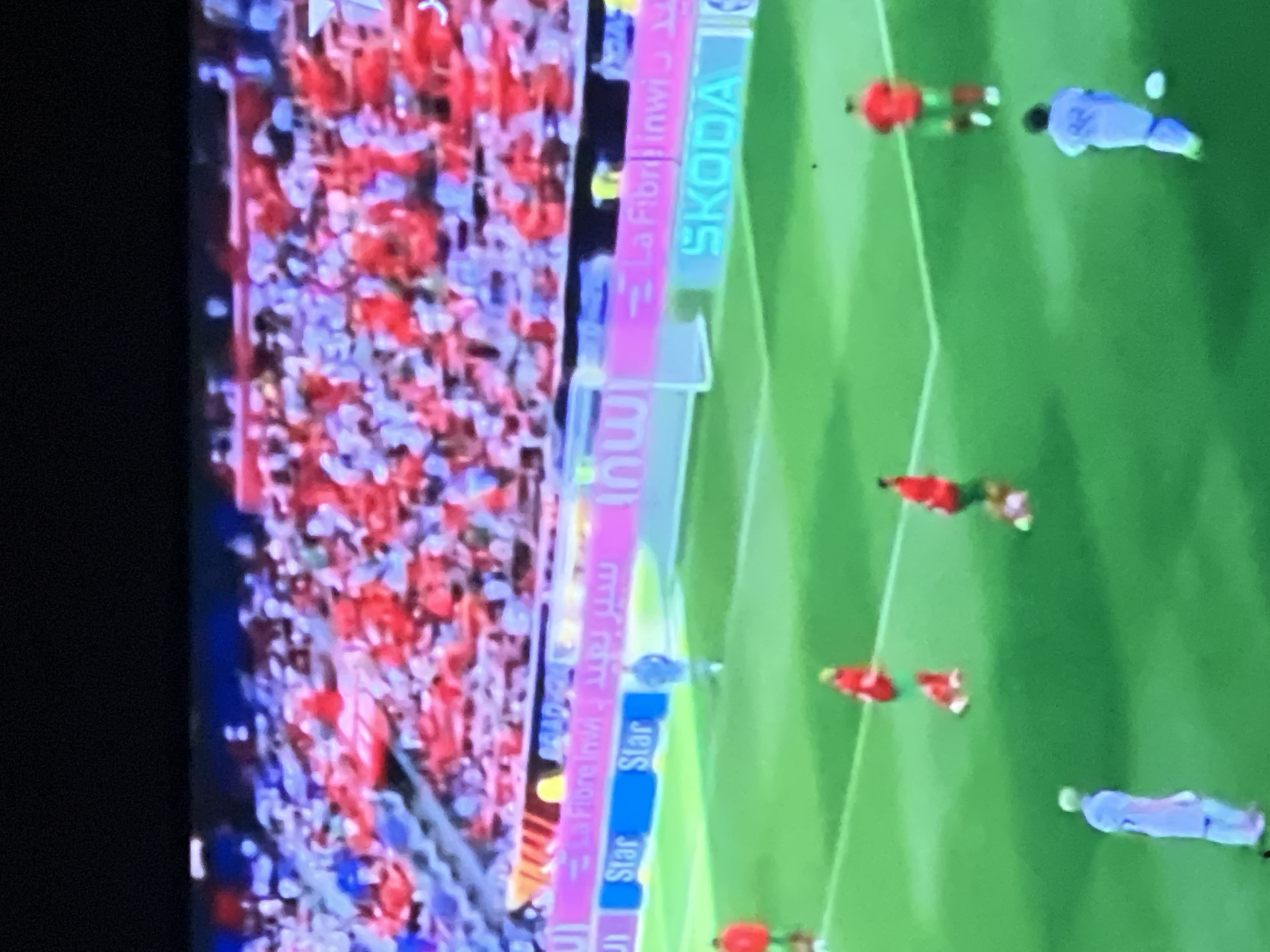 The image captures a football (soccer) match in progress, likely at a stadium in Casablanca, Morocco, indicated by the prominent "inwi" and "La Fibre inwi" advertisements visible on the LED hoardings, along with "SKODA" and "Star," displayed in both Arabic and Latin scripts.
The scene focuses on the green grass pitch where several players are active. One team wears red jerseys and green shorts, with at least three players visible on the right and center of the field; one player in red is on the ground near the center. Another player, likely from an opposing team or a goalkeeper, is dressed in light blue or white, positioned in the foreground and appearing to control a white football. Another player in light blue is walking towards the foreground, and a figure in light blue is standing near the goal area on the left side of the frame, possibly an official or a substitute player. The goal itself is partially visible.
In the background, tiered stadium seating is densely packed with spectators, creating a mosaic of red, white, and other colors, suggesting a lively and well-attended event. The upper portion of the image shows the dark stadium roof or sky. The overall lighting is bright, illuminating the pitch and the crowd.