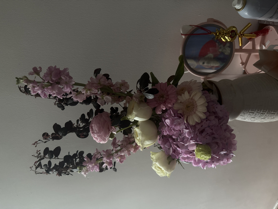 This image, taken in Ngũ Hành Sơn Ward, Vietnam, depicts a static indoor scene. A large, ornate floral arrangement is centrally positioned in a light-colored ceramic vase with a textured, possibly ribbed pattern. The bouquet comprises various flowers and foliage: tall stems of light pink/lavender stock flowers, dark leafy branches, a single light pink ranunculus, multiple creamy white roses, a mass of purple hydrangeas, and several light pink gerbera daisies. A single pale green flower bud is visible within the hydrangeas. The background is a plain, light-colored wall. To the right of the floral arrangement, a small, pink, cat-ear shaped vanity mirror is present, reflecting a blurred interior scene possibly including a light-colored object and a red element. A gold-colored ornamental charm, shaped like a money bag with a "福" (Fú) character and a red tassel, hangs from the mirror's stand. Partially visible on the far right is a light blue aerosol can or bottle. No individuals or active interactions are observed. The scene is illuminated by ambient light, casting soft shadows.