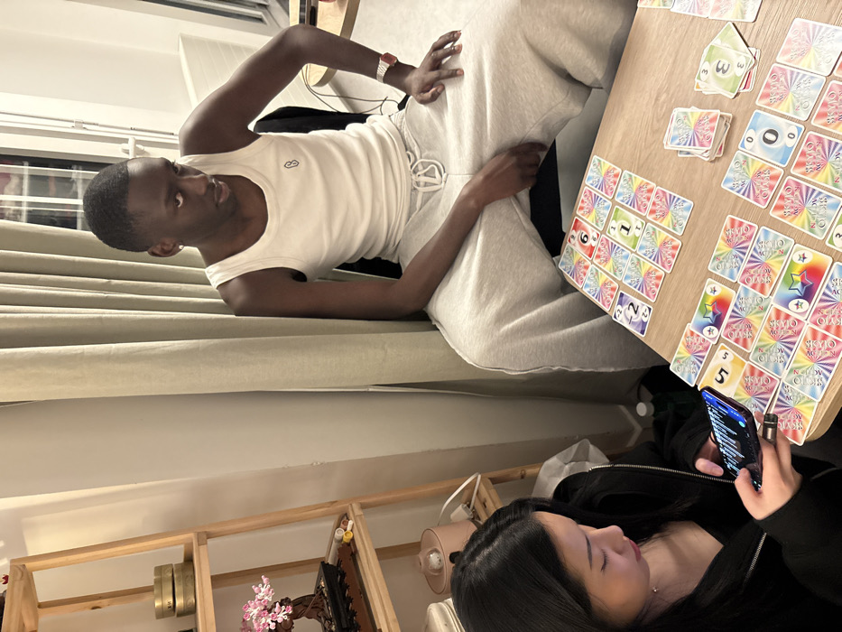 The scene depicts an indoor, residential setting with two individuals engaged around a light wooden table. The specific location within Paris, France, is not discernible from the image.
In the foreground, a female with long dark hair, wearing a black long-sleeved top, is seated. She is intently focused on a dark-colored smartphone held in both hands, its screen illuminated with visible text or icons.
To her right, partially in the background, a male with short dark hair and dark complexion is seated. He wears a white ribbed tank top with a small dark logo on the left chest and light grey drawstring sweatpants. A red-strapped digital watch is visible on his left wrist, and a silver stud earring in his left ear. He is looking downwards towards the table with a contemplative expression.
The wooden table between them is covered with colorful cards, arranged in various spreads and small stacks, indicating an ongoing card game. The cards feature radial rainbow patterns with numbers (0, 1, 2, 3, 5, 6) and star symbols.
To the left of the female, a light wooden shelving unit stands against a plain white wall. On its top shelf are two stacked brass-colored cylindrical containers. The shelf below holds a pink decorative bonsai tree model on a dark wooden stand. The third shelf down displays a pink cylindrical device with a white power cord, possibly a humidifier or diffuser.
Behind the male, light beige curtains hang, partially revealing a window or balcony door frame and a white radiator on the far right. The room is illuminated by artificial light, casting soft shadows.