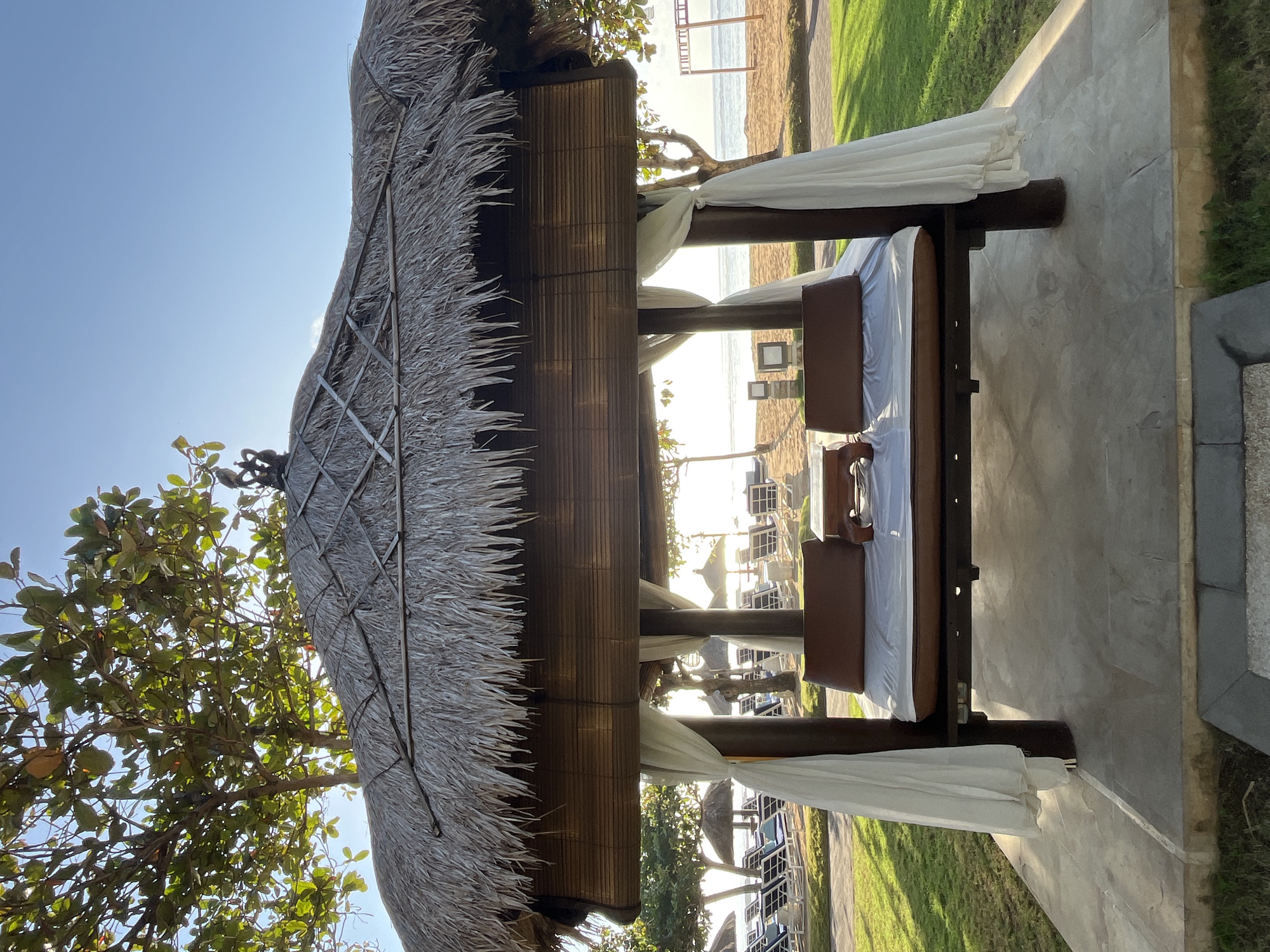 A daybed with a thatched roof is situated on a paved area overlooking a beach. The daybed has a white mattress and two brown pillows, with white curtains hanging from its posts. In the background, beach chairs and umbrellas are visible on the sand, with the ocean beyond. Trees line the area between the daybed and the beach. The scene appears to be in Kuta Selatan, Indonesia, known for its beautiful beaches and resorts. The sun is setting, casting a warm glow over the scene.