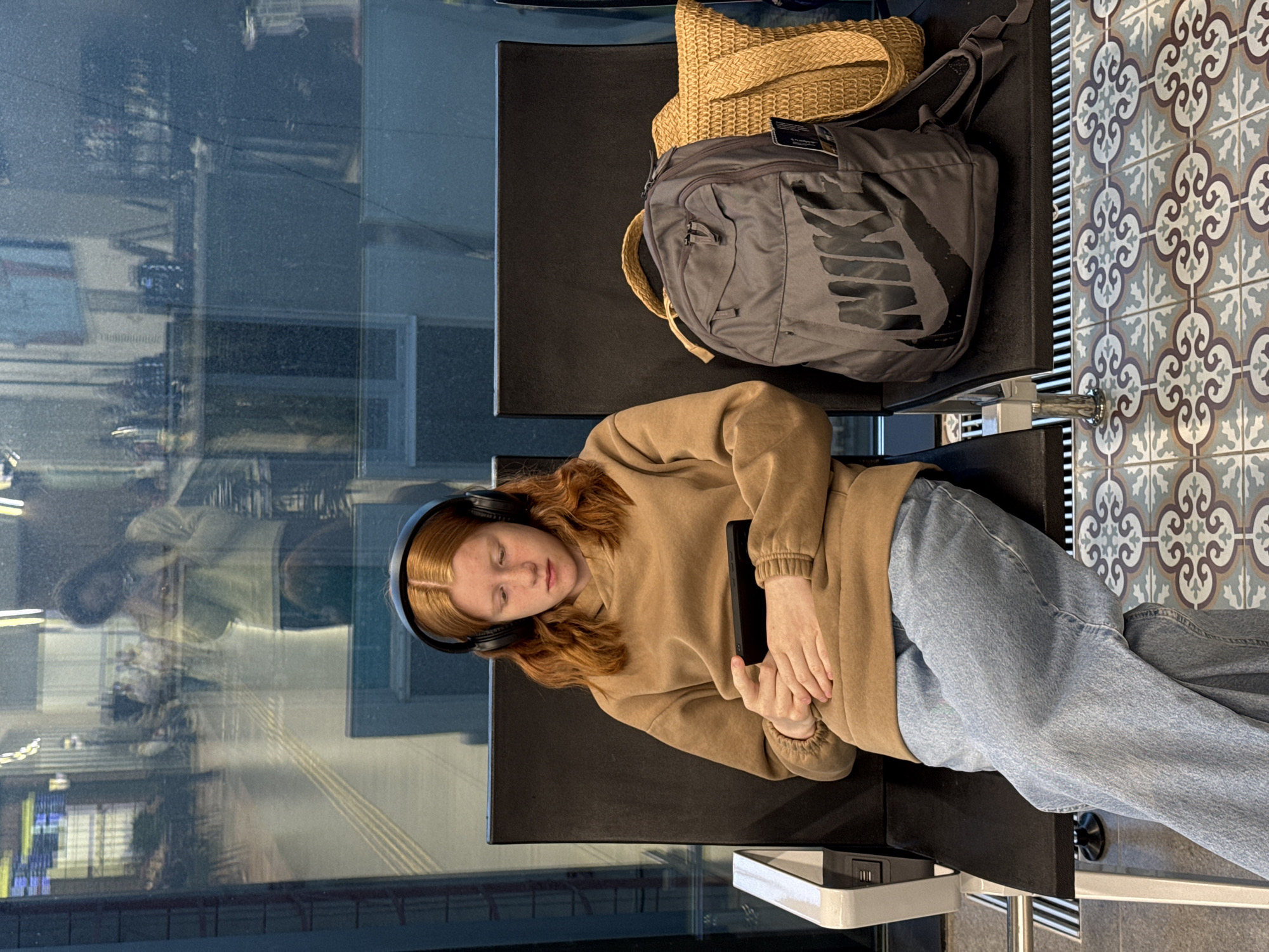 The image captures a young person resting on a black public bench, likely in a waiting area such as an airport or train station. The person, appearing to be a young female with reddish-blonde hair, is lying on her side with eyes closed, suggesting she is sleeping or deeply resting. She wears black over-ear headphones, a light brown hooded sweatshirt, and light blue jeans. A black smartphone or small tablet is held in her clasped hands, resting on her stomach.
To her upper right, on an adjacent seat, are two bags: a light brown woven straw bag and a grey backpack with a prominent, stylized "NIKE" logo visible on its side in a darker grey. The floor to the right of the seating features decorative patterned tiles in shades of grey, white, and brown/blue. A large window occupies the left side of the frame, showcasing blurry reflections of an indoor environment, including what appears to be a distorted reflection of another person or structure. A white charging module with USB ports is attached to the seating structure beneath her feet. The lighting suggests it is daytime. The scene conveys an atmosphere of quiet waiting or travel.