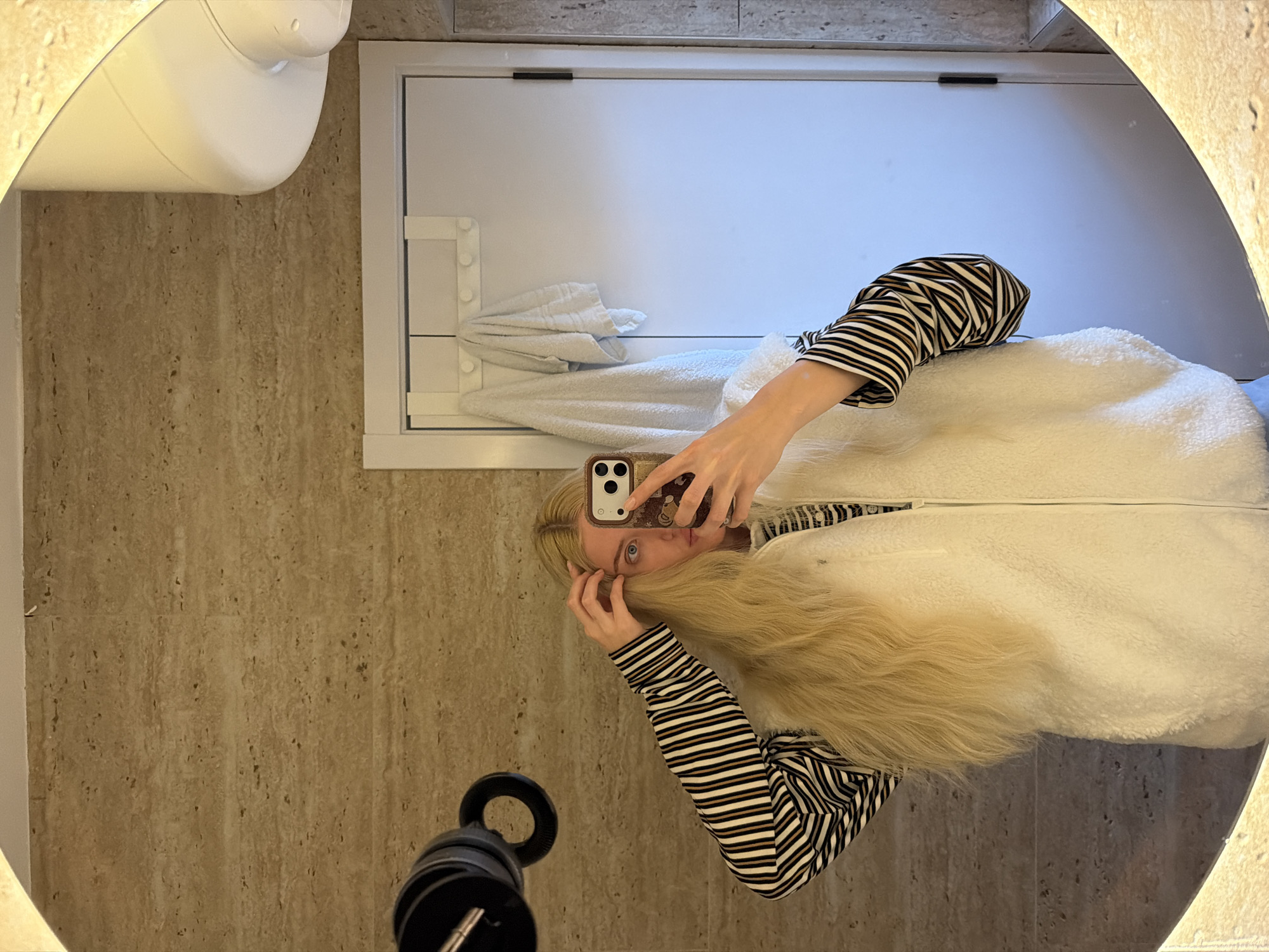 A blonde woman is taking a selfie in a bathroom mirror. She is wearing a striped shirt and a white vest. The bathroom has beige tile walls and a white cabinet. There is a toilet to the left of the cabinet. The location is unknown and there is no evidence of it being in the city of Belgrade, Serbia.