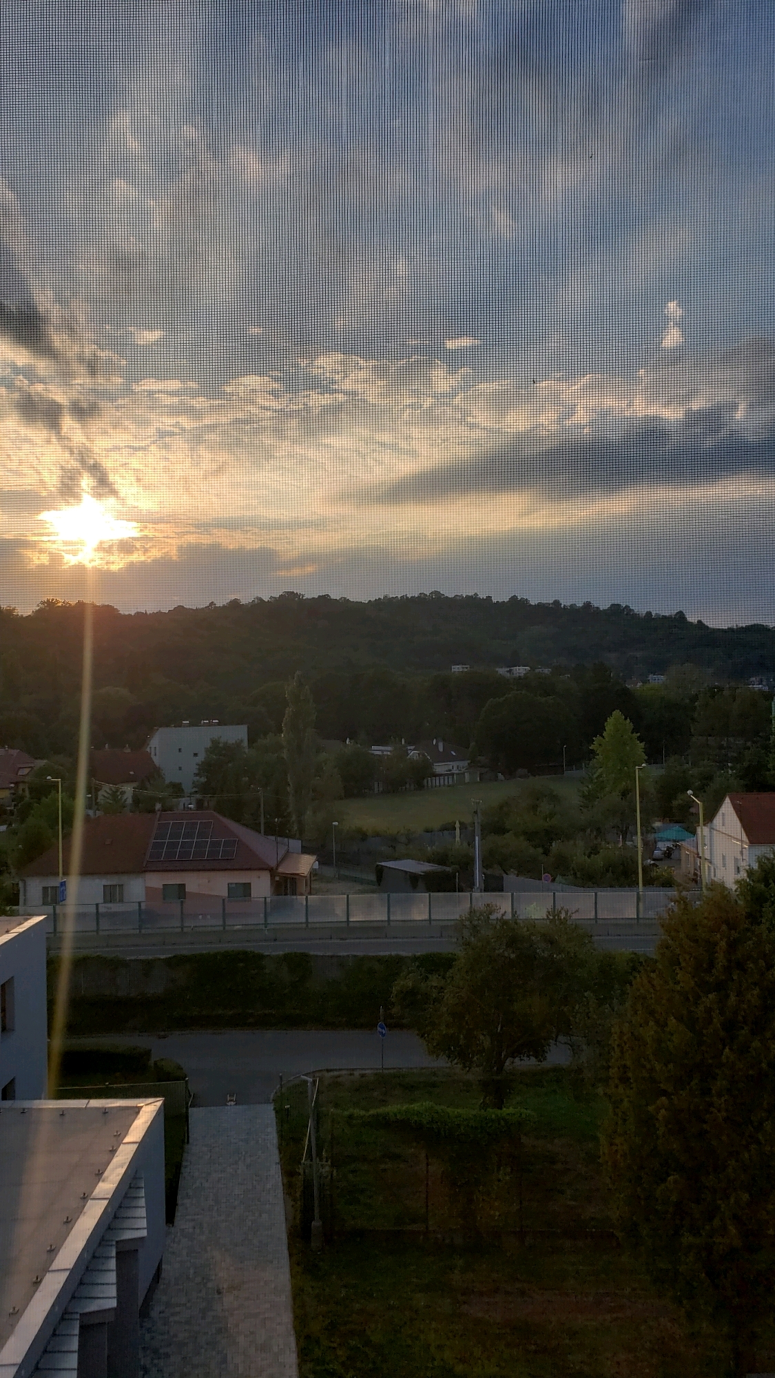 A sunset view from a window with a screen is depicted. The sun is low on the horizon, casting a warm glow and creating light rays. Clouds are scattered across the sky, with some appearing wispy and others denser. Below the sky, a tree-covered hill forms the backdrop. In the middle ground, residential buildings with red roofs and a modern white building are visible. A road runs horizontally across the image, with a fence along its side. The foreground shows part of a modern building and a paved walkway. The location is identified as Prešov, Slovakia.