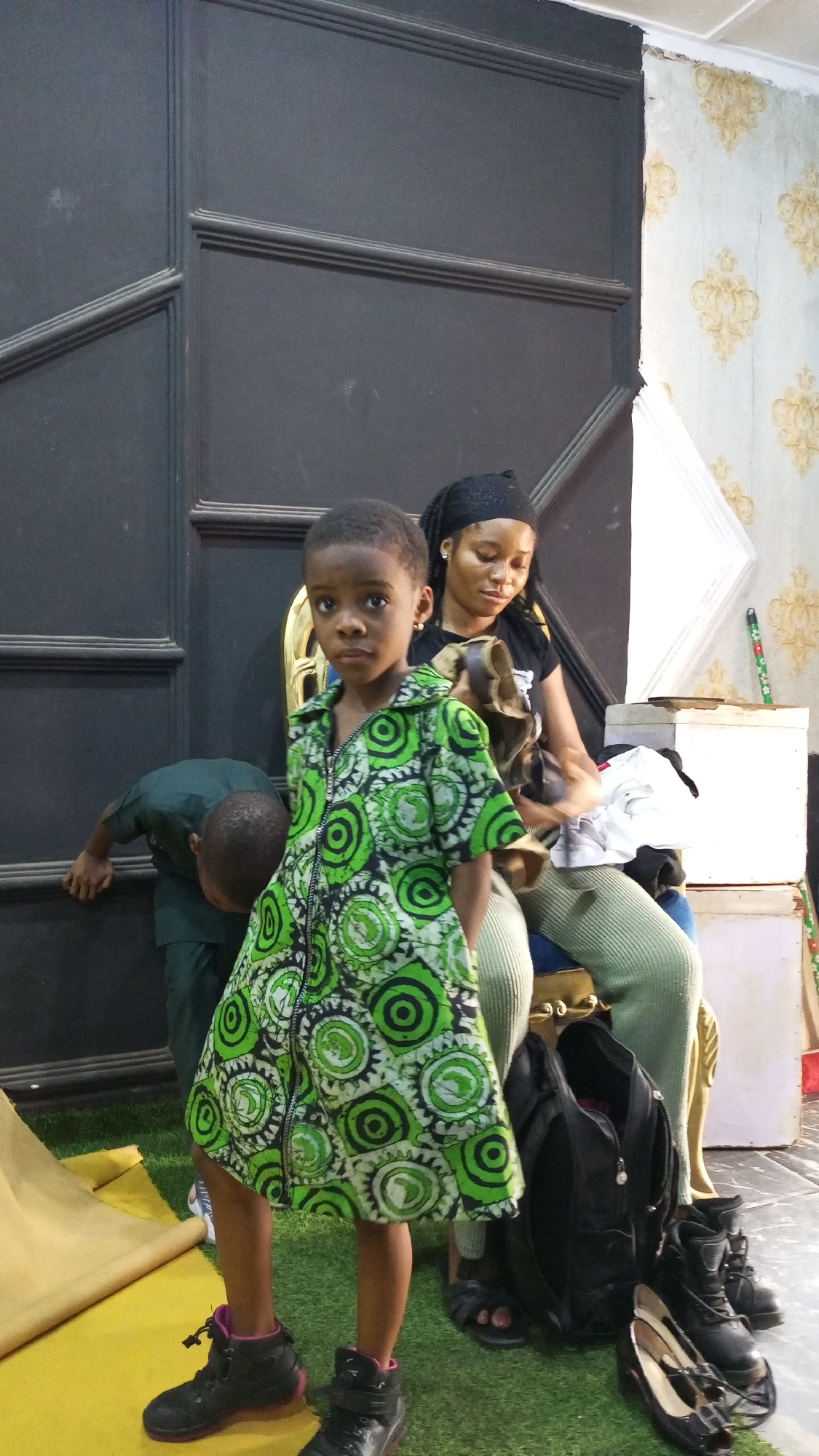 The image shows a young child wearing a green and black patterned dress and black shoes, standing in front of a dark door. Behind the child, a woman is seated on a gold-colored chair, wearing a black headscarf and a black t-shirt. Another child is bending over near the door. A black backpack and a pair of black shoes are visible on the floor next to the chair. The background includes a wall with patterned wallpaper and a white box. The setting appears to be indoors, possibly in a room or studio, with elements suggesting a staged or arranged environment, such as artificial grass and a yellow sheet on the floor. The location in the city of Unknown, Unknown, cannot be determined from the image.