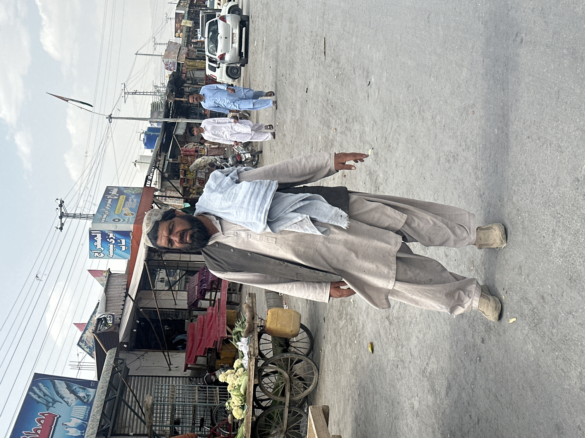 In Quetta City Tehsil, Pakistan, a man with a beard and wearing traditional Pakistani clothing stands next to a vegetable cart, seemingly discarding something from his hand. The cart contains cauliflower and a yellow container. In the background, two men in white and light blue shalwar kameez outfits walk along a street lined with shops under a cloudy sky. Cars are parked along the road. Utility poles and wires crisscross overhead.