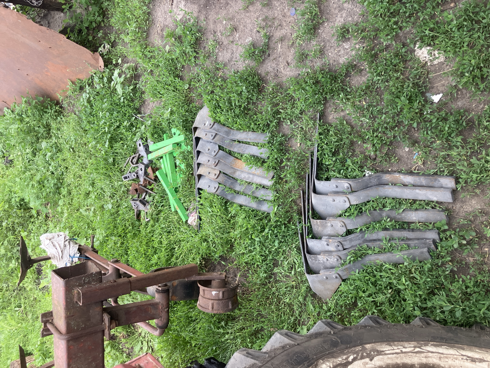 This image depicts an outdoor scene with agricultural equipment and vegetation. In the foreground, the tire of a large tractor is visible on the left. To the right of the tire, several metal plowshares are arranged in a row, lying on the ground amidst green grass and dirt. Behind the plowshares, more metal parts, some green plastic components, and other miscellaneous metal pieces are scattered on the ground. Further in the background, a red and rusty piece of farm machinery, possibly a cultivator or plow frame, is positioned on the left. A large, rusty sheet of metal leans against the background on the right. The ground is covered with grass and patches of bare earth. The environment appears to be a rural or farm setting, likely in the city of Vovkivka, Ukraine, given the provided location context. There are no people or active events depicted.