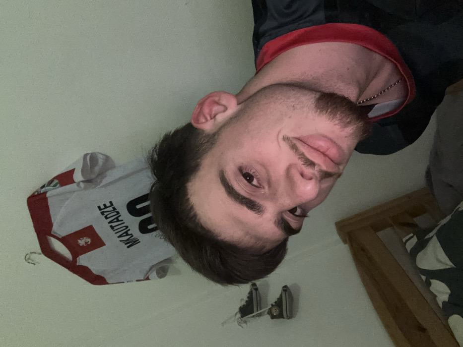 A man with dark hair and a mustache is facing forward in a slightly tilted image. He wears a dark shirt with a red collar and a chain necklace. Behind him on the wall hangs a white soccer jersey with red accents and the name "Mkautadze" and the number "00" printed on it. There is also a pair of black shoes on the wall, and a wooden bed frame can be seen in the background.