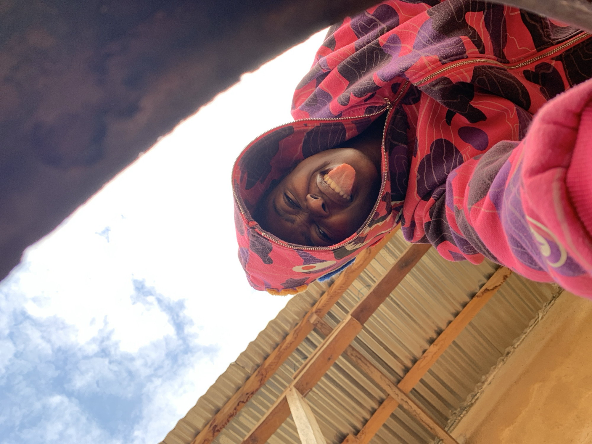 The image displays a single young individual, likely an adolescent, positioned centrally and viewed from a low-angle perspective in Rafin Par, Nigeria. The person has dark skin and is wearing a bright pink, red, and purple camouflage-patterned hooded sweatshirt, with the hood up. A yellow knit element is visible under the hood on the left side of their head. The individual is looking directly at the camera, making a playful expression by scrunching their face and extending their tongue. The background, from left to right, reveals a segment of blue sky with scattered white clouds above a corrugated metal roof supported by exposed wooden beams. A textured, light brown wall is visible below the roof structure on the left. A dark, out-of-focus object partially obstructs the upper right portion of the frame. The scene is well-lit, indicating daylight conditions.