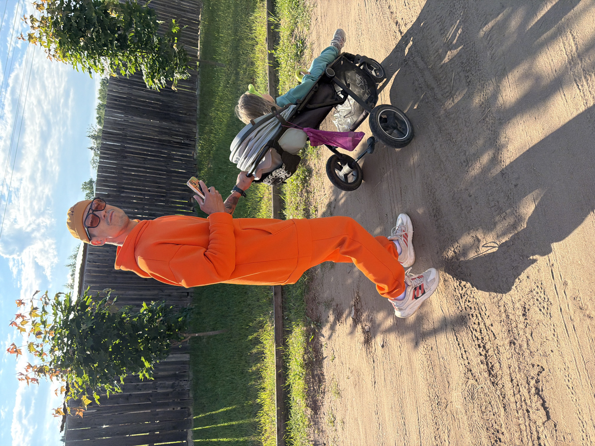 An outdoor scene captures a person pushing a stroller on a sunny day in what appears to be Chaikovsky, Russia. The image is oriented vertically but depicts a horizontal perspective.
In the foreground, an adult, dressed in a bright orange tracksuit and a beige beanie, walks along a dirt or gravel path. They wear dark sunglasses with orange frames and white sneakers with red accents. On their left wrist is a dark watch, and tattoos are visible on their left forearm. The person holds a smartphone with a colorful case in their right hand, seemingly looking at the screen.
Behind the adult, a child is seated in a dark-colored stroller. The child wears light blue clothing and appears to be looking away or resting. Various items, including a grey canopy or cover and a purple bag, are attached to or stored in the stroller.
The path shows tire tracks and footprints. To the left of the path, a dark wooden fence runs parallel, with green grass and several trees visible beyond it. The sky above is bright blue with scattered white clouds. Long, distinct shadows on the ground suggest it is either late afternoon or early morning. The overall atmosphere is peaceful and casual, depicting a leisurely outdoor activity. No legible text is visible in the image.