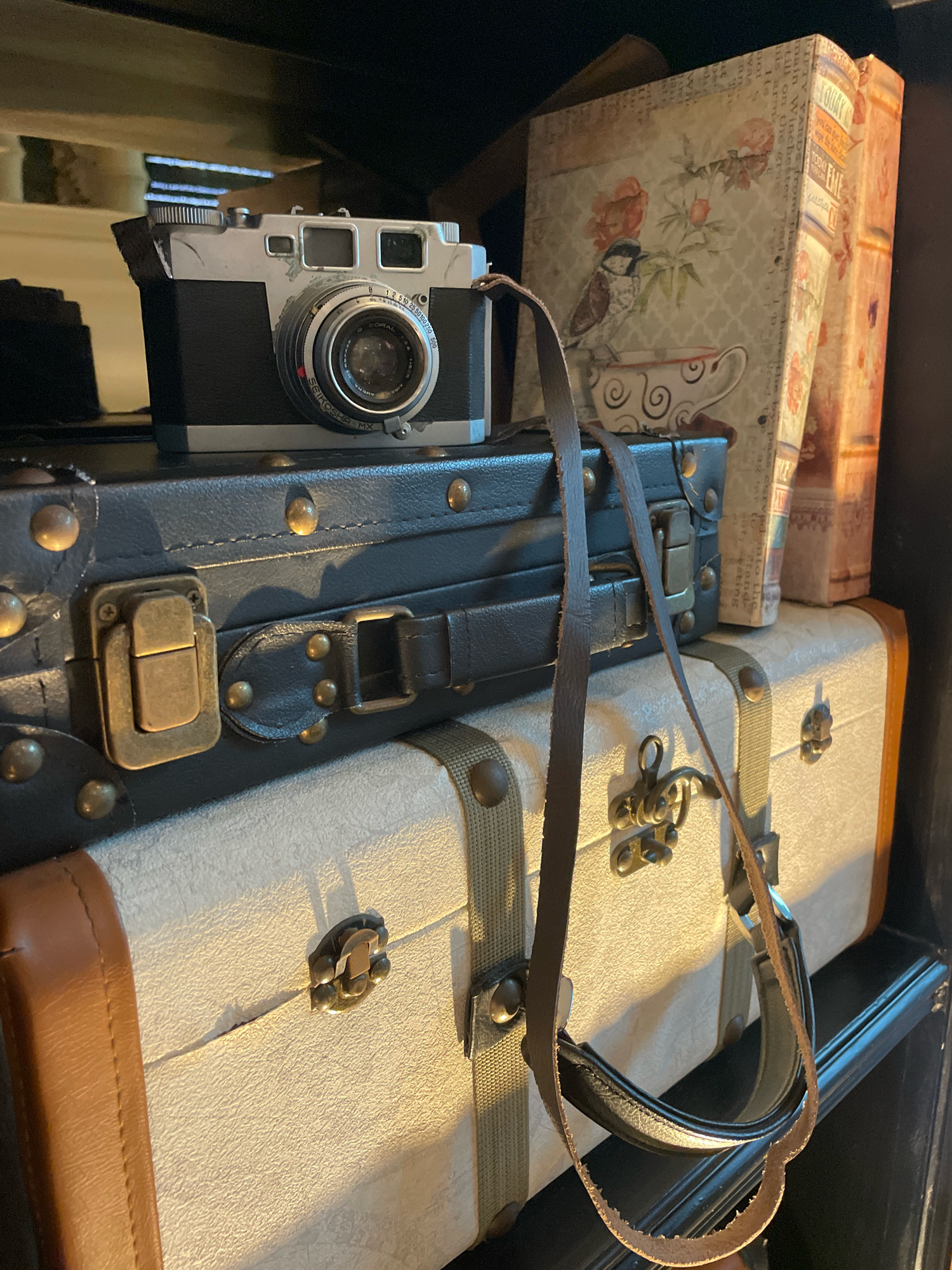 The image shows a stack of vintage-style suitcases in an indoor setting. The top suitcase is black with brass-colored hardware and a leather strap. A vintage camera sits on top of this suitcase. The middle suitcase is off-white with tan trim and brass-colored hardware. The bottom suitcase is also off-white with tan trim and brass-colored hardware. Behind the suitcases, two decorative books are visible. The lighting suggests an indoor environment, possibly a home or shop. There is no indication of the specific location within Pattaya City, Thailand.
