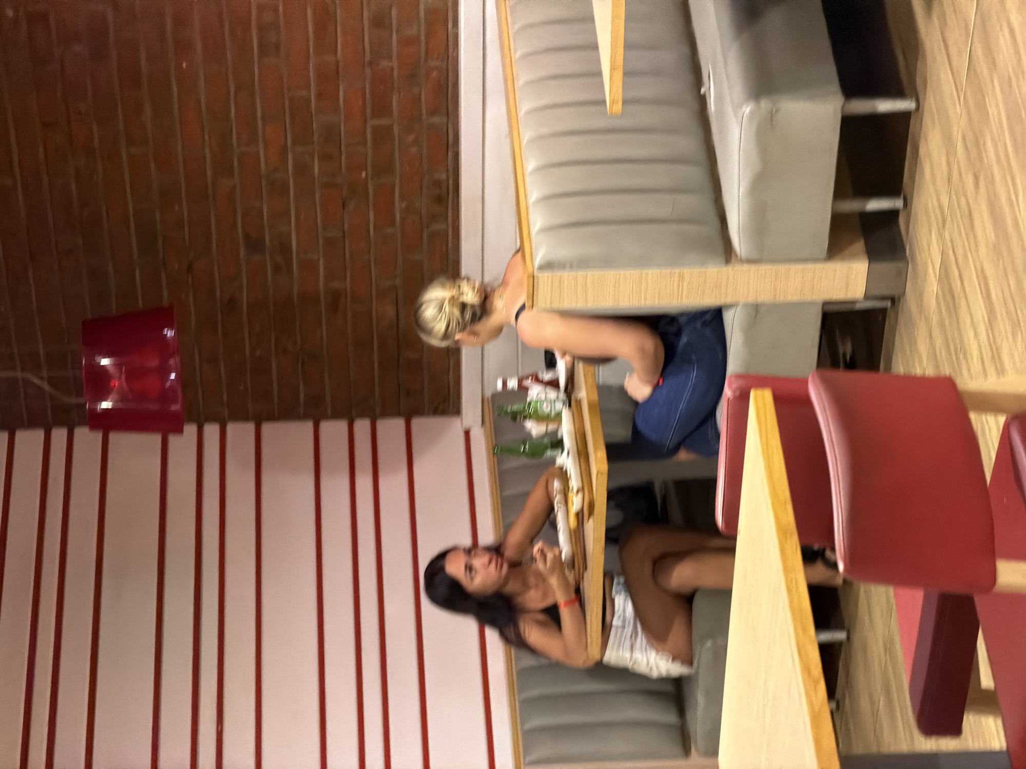 Two young women are seated in a restaurant booth in Sharm El Sheikh, Egypt. The woman on the left has long dark hair and is wearing a black tank top and white shorts. She is looking down at her meal. The woman on the right has blonde hair pulled back and is wearing a dark tank top and blue jeans. She is facing away from the camera. The restaurant has a brick wall on the right and a white wall with red stripes on the left. A red pendant light hangs from the ceiling. There are wooden tables and red chairs in the foreground.