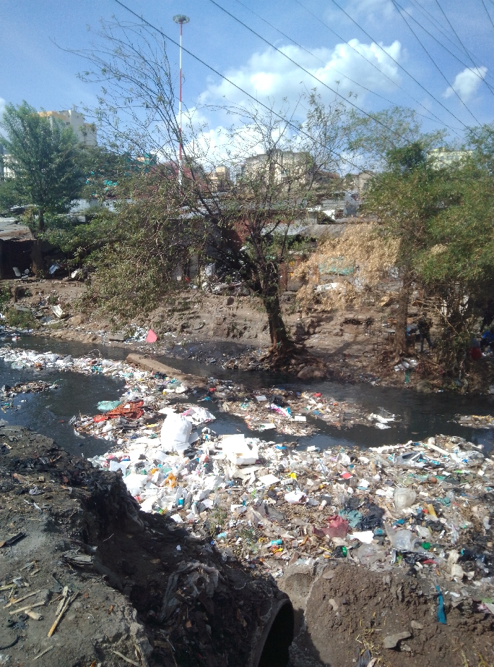 The image displays a severely polluted waterway in an urban informal settlement of Nairobi, Kenya, with a prominent red and white striped telecommunication mast visible in the upper-left background, indicative of areas near the Globe Roundabout or Mathare. The dark, murky water of the river or stream is almost entirely obscured by a dense layer of solid waste, predominantly composed of plastic bags, plastic bottles, and white polystyrene foam, forming extensive floating mats.
The riverbanks are steep, eroded, and muddy, also heavily laden with refuse. In the immediate foreground, a trench of dark earth leads to a partially exposed concrete pipe, suggesting a discharge point into the polluted water. On the far bank, informal structures made of corrugated metal and other makeshift materials are visible among trees with sparse foliage and patches of dry vegetation. Beyond these structures, multi-story concrete buildings typical of urban residential areas are discernible.
Two indistinct human figures are visible on the far right bank: one stands near a tree, partially obscured, and another is further to the right near a blue object. Overhead, several dark utility lines crisscross against a partly cloudy blue sky. The scene depicts significant environmental degradation characterized by extensive waste accumulation in the waterway and surrounding area.