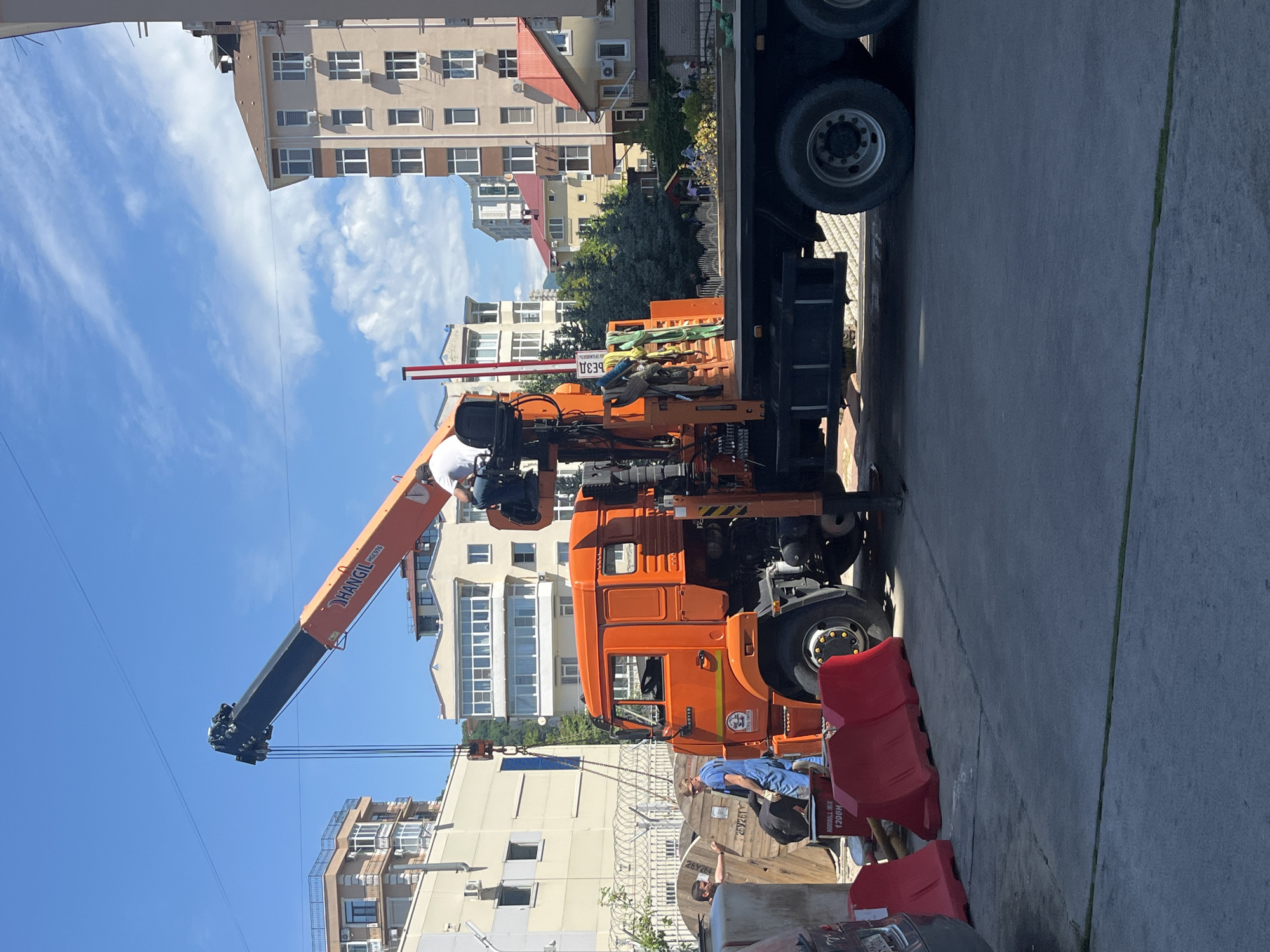 An orange crane truck is positioned on a paved surface in what appears to be an urban or semi-urban setting in Sochi, Russia, under a bright blue sky with scattered clouds. The truck features a prominent orange and black telescopic crane arm, which extends towards the left side of the frame. One man, wearing a white shirt and hat, is visible in the crane's operator area, seemingly overseeing or engaging with the equipment.
Nearby, a second man in a blue shirt is observed interacting with large wooden spools, which are likely used for holding cables or wires. These spools bear various alphanumeric markings, including "2BY2G1", "1200M", and the brand names "KOBELL MIX" and "HORDILL MIX". One spool also shows "СБЭ30" and another "3033S". Red plastic barriers are also visible on the ground.
The background features multi-story residential or commercial buildings with balconies and air conditioning units, characteristic of a developed city. Green trees and other foliage are interspersed among the structures. The overall scene suggests ongoing construction, maintenance, or installation work, likely related to utilities or infrastructure given the presence of the crane and cable spools.
Visible text on the crane arm includes "HANGLI" and "HC3VFA". The truck's cab door has a logo and text, partially visible, which seems to indicate its affiliation with a municipal or utility service, possibly "ГОРОДСКИЕ СЕТИ" (City Networks). The time of day appears to be bright daylight, suggesting a clear and sunny day.