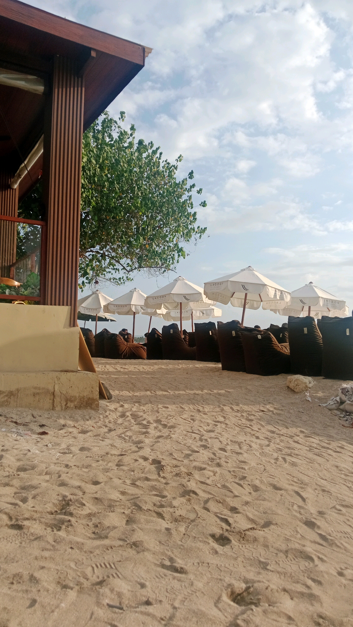 The image depicts a beachside seating area, likely part of a resort or cafe in Kuta, Indonesia. The foreground is dominated by textured sand with numerous footprints. To the left, a partially visible building with vertical wooden slats and a brown roof is present. In the midground, a row of white umbrellas with the word "LAGUNA" visible on them stands over brown beanbag chairs. The beanbags appear to be unoccupied, with one person visible in the distance sitting on one. Behind the seating area, a lush green tree provides shade. The background features a bright, cloudy sky, suggesting daytime. The scene is serene and suggestive of a relaxing atmosphere.