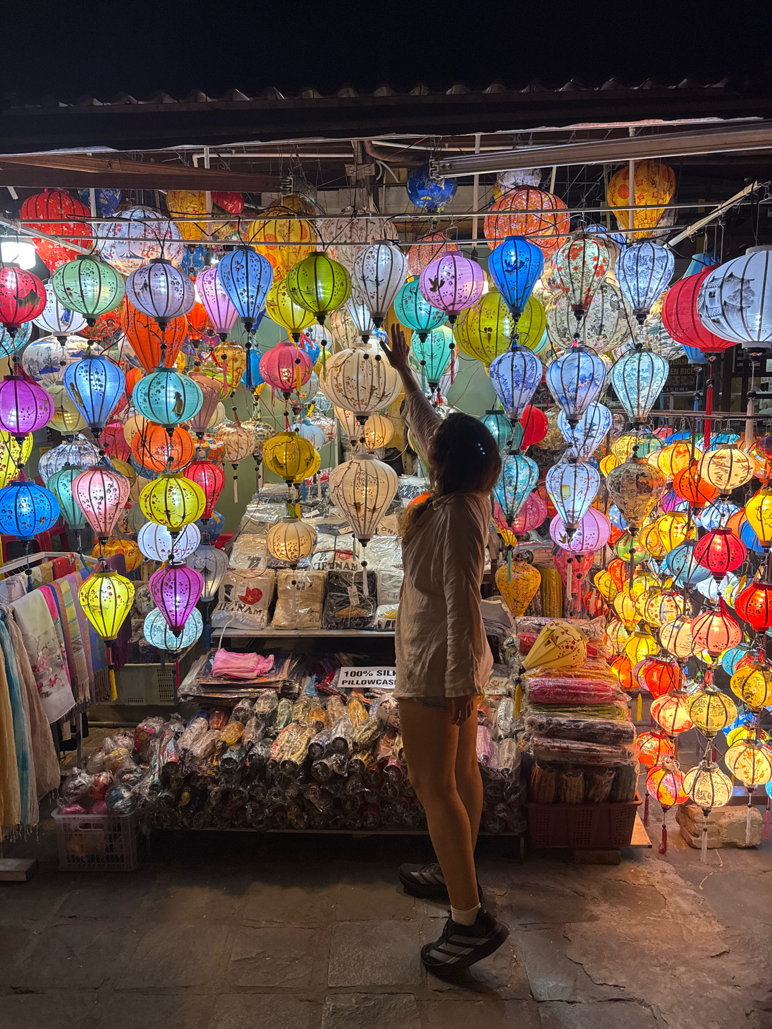 This image captures a nighttime scene in the ancient town of Hoi An, Vietnam, likely on a street lined with shops. The focal point is a stall overflowing with a vibrant array of colorful lanterns, illuminated and hanging from overhead structures. The lanterns are of various shapes and sizes, predominantly round and teardrop, decorated with intricate patterns and designs.
A lone individual, seen from the back, stands in front of the lantern display, reaching up towards one of the hanging lanterns. The person is wearing a light-colored shirt and shorts, and dark athletic shoes. Their posture suggests they are interacting with the lanterns, possibly admiring or selecting one.
The stall also displays various other items for sale, including textiles, such as scarves or fabric rolls, neatly folded and stacked. Some items are packaged, indicating they are ready for purchase. The ground in front of the stall appears to be paved with stone, showing signs of wear and the texture of age. The overall atmosphere is illuminated by the warm glow of the lanterns, creating a festive and inviting ambiance characteristic of Hoi An's night markets.