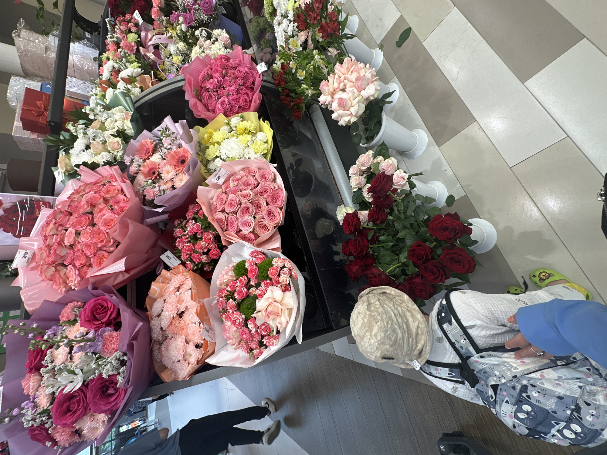 The image shows a variety of bouquets displayed on a black shelving unit, likely in a flower shop or market. The bouquets vary in color and flower type, including roses, carnations, and mixed arrangements in shades of pink, red, white, and yellow. Some bouquets are wrapped in paper or cellophane.
To the right of the flowers, there is a patterned floor with diagonal stripes in varying shades of gray. An individual in blue clothing is present, possibly a customer or employee interacting with the flower display. A shopping cart is also partially visible. A person's legs and feet are visible near the floor.
It is impossible to confirm the location in Nizhny Novgorod, Russia, based solely on visual evidence.