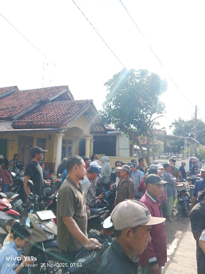 A crowd gathers outside a single-story house with a red-tiled roof. Many individuals are on motorcycles, while others stand around, observing. Most of the people are men, with some wearing hats. The scene is filled with vehicles, and a tree stands in the background, casting shadows. The photograph is taken on a sunny day, with the sun visible in the sky.