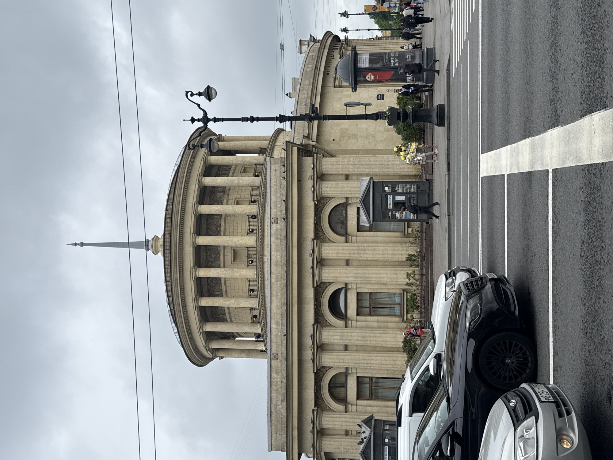 The image shows a street scene in Saint Petersburg, Russia, featuring an architectural structure with a prominent rotunda supported by columns, topped with a spire. This building is likely the Eliseyev Emporium, located on Nevsky Prospekt. The sky is overcast. In the foreground, there are parked cars. People are walking on the sidewalk in the background. A pedestrian crosswalk is visible on the street. A lamppost stands near the building. A newsstand or kiosk is on the sidewalk.