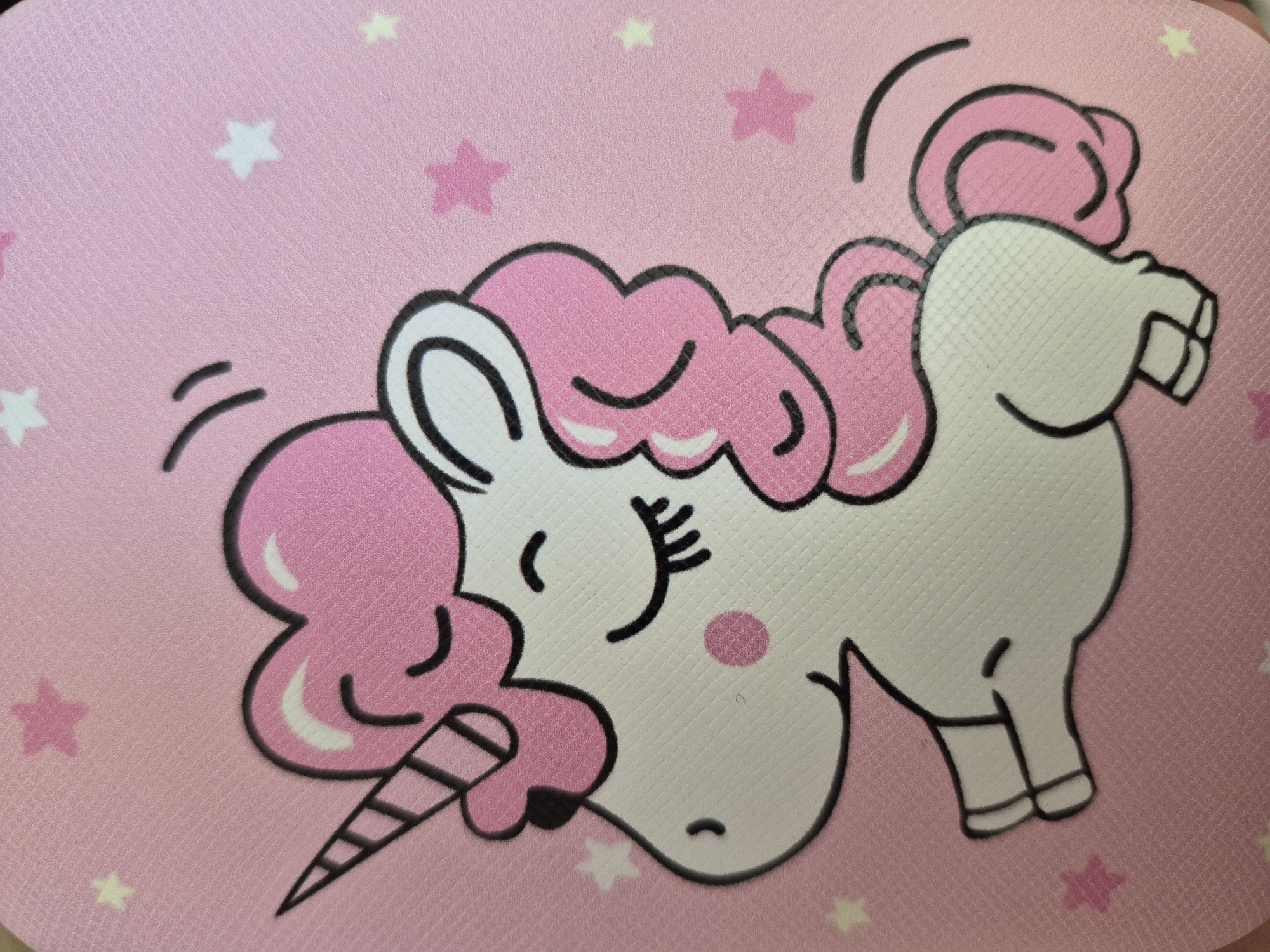 The media file displays a close-up image of a vibrant, cartoonish unicorn against a pink background. The unicorn is predominantly white with a fluffy pink mane and tail, outlined in black. Its horn is depicted with black and white stripes. The unicorn has a cute, smiling expression with closed eyes and eyelashes, a small pink blush on its cheek, and a tiny, subtle smile on its snout. Curved lines above its head and near its tail suggest a dynamic, possibly playful, motion. The background is a textured light pink, patterned with scattered stars in both lighter yellow and darker pink hues. The overall aesthetic is whimsical and childlike. No text is visible in the image.