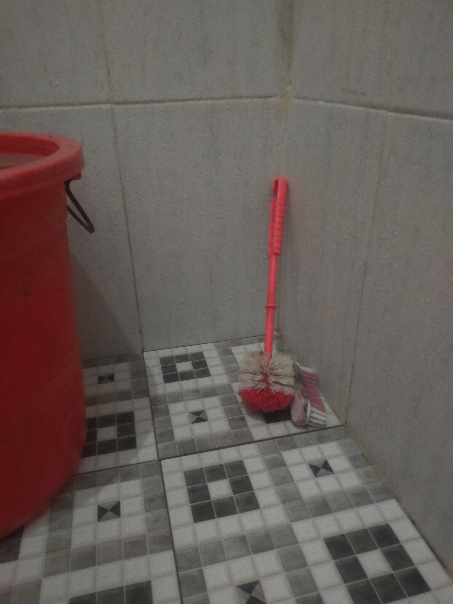 A toilet brush with a red handle and white bristles stands upright on a tiled floor in the corner of a room. To the left, a red plastic bucket is partially visible. The walls are tiled with small, light-colored squares, and the floor is also tiled with a geometric pattern of light and dark gray squares. The scene appears to be in a bathroom or utility area. The location in Tegalandong, Indonesia is not evident from the image itself.