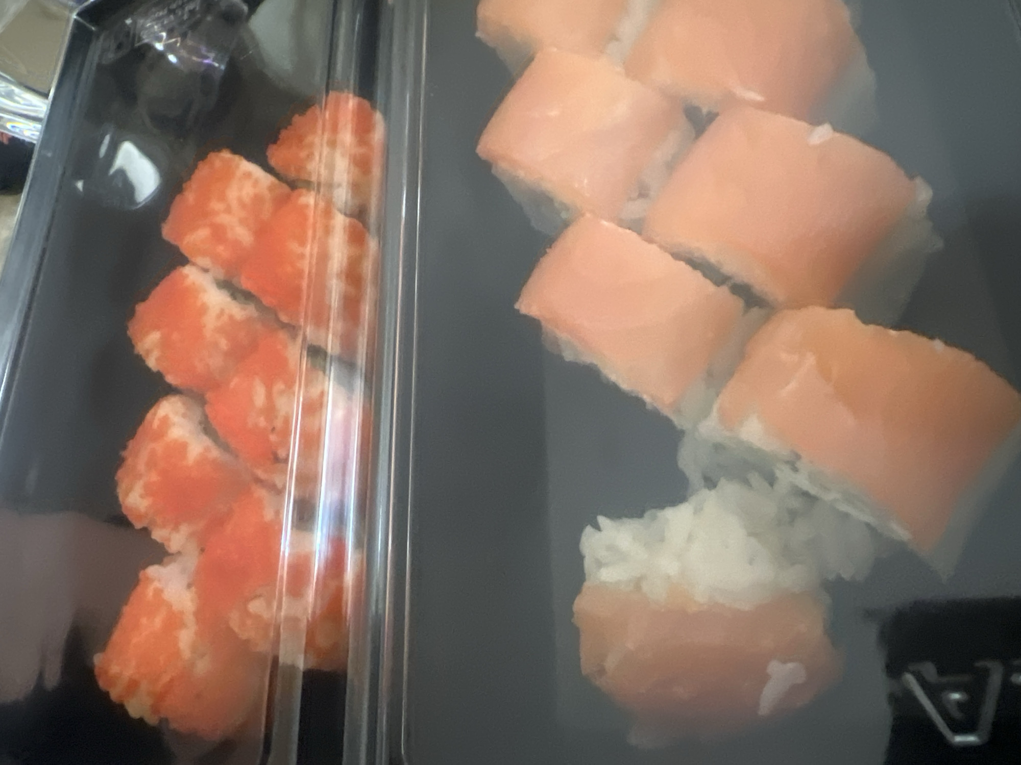 The image shows two rows of sushi in plastic containers. The top container has several pieces of sushi that appear to be California rolls, with orange roe on the outside and white rice on the inside. The bottom container has several pieces of sushi that appear to be salmon nigiri, with slices of salmon on top of rice. The sushi is arranged in a linear fashion within each container. The lighting suggests it is indoors. The location is unknown.