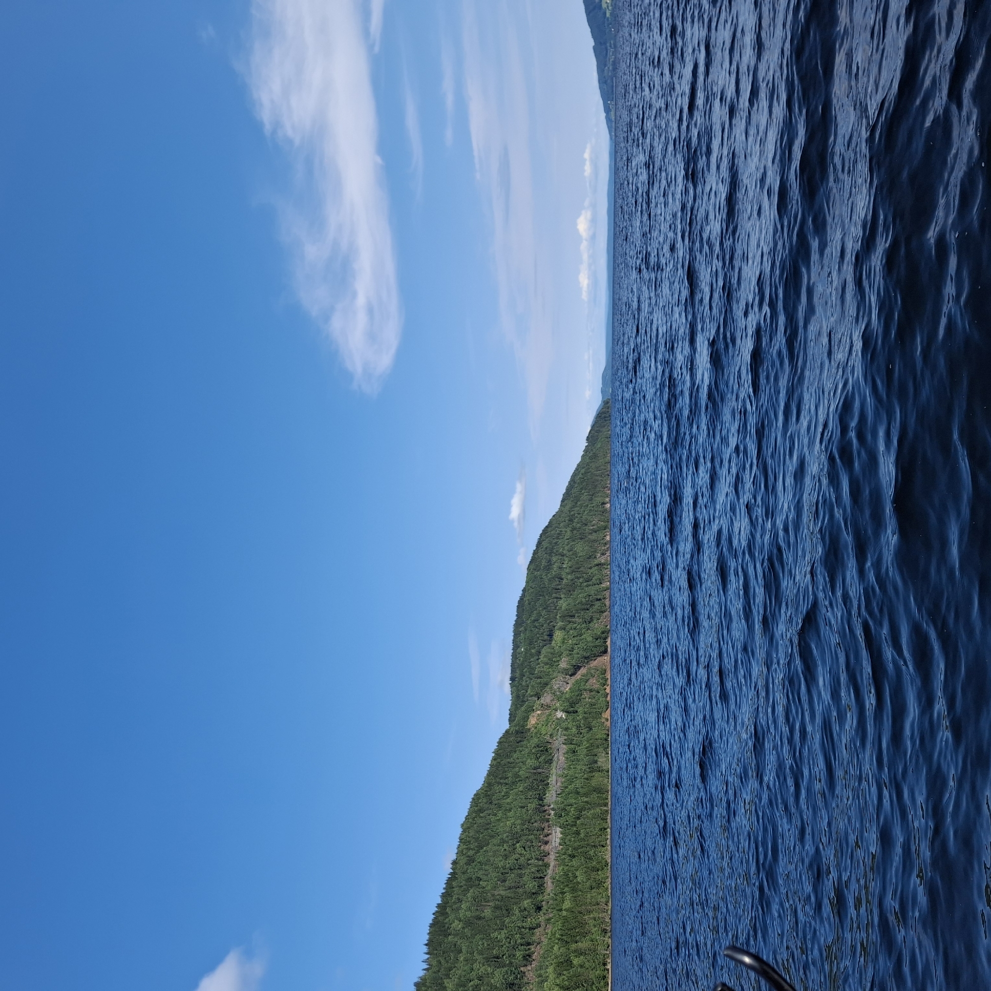The image shows a serene natural landscape on a clear day, likely in the vicinity of Minnesund, Norway. The scene is dominated by a vast expanse of deep blue water, reflecting the bright blue sky above. Gentle ripples on the water's surface suggest a calm day.
The left side of the image features a densely forested hill that slopes down to meet the water's edge. The trees are predominantly green, indicating a healthy forest. A road or path is visible cutting through the trees on the hillside, suggesting human presence or access. The terrain appears rugged and natural.
In the distance, the water stretches towards a horizon where more landmasses and hills can be seen, also covered in vegetation. The sky is a vibrant blue with a few wispy white clouds scattered across it, adding to the picturesque quality of the scene. The overall impression is one of natural beauty and tranquility.
