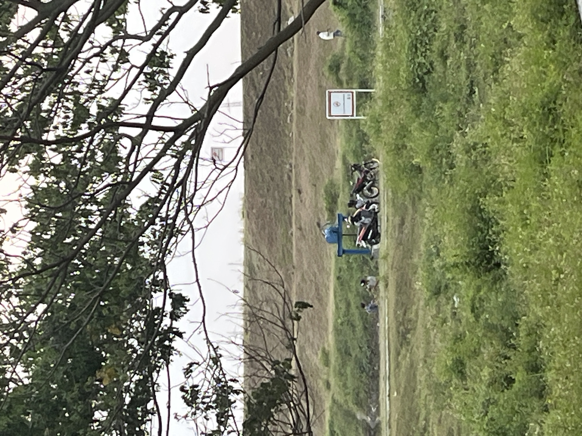 The image depicts an outdoor scene with a large tree in the foreground, its branches partially obscuring the view of the background. The middle ground features a grassy area with several people and motorcycles. A blue structure, possibly part of a water pump or filtration system, is visible near the motorcycles. Further in the background, there's a raised embankment covered in dry grass. A white sign with a red symbol and text is positioned on the right side of the embankment. Another person in a white shirt is walking away in the distance on the right. The overall environment suggests a rural or semi-rural area, possibly near a waterway or agricultural land. The lighting indicates it might be overcast or late afternoon. The specific location in Besuki, Indonesia, is suggested to be an area with some form of water management infrastructure.