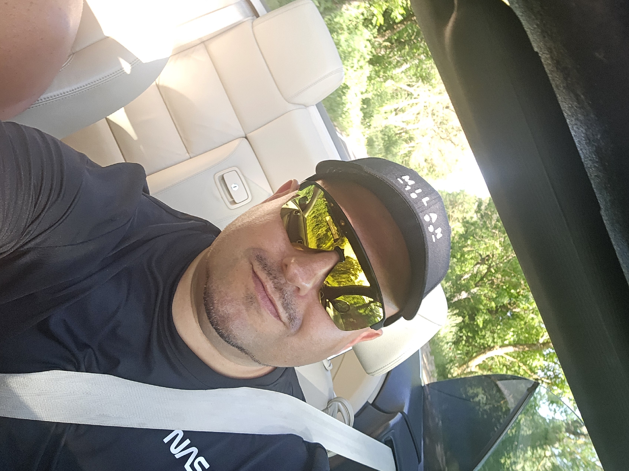 A man is sitting in a convertible, wearing a black baseball cap and sunglasses. He has a seatbelt on. The car interior is light-colored. Green trees are visible in the reflection of his sunglasses and through the car window. The location is not discernible from the image.