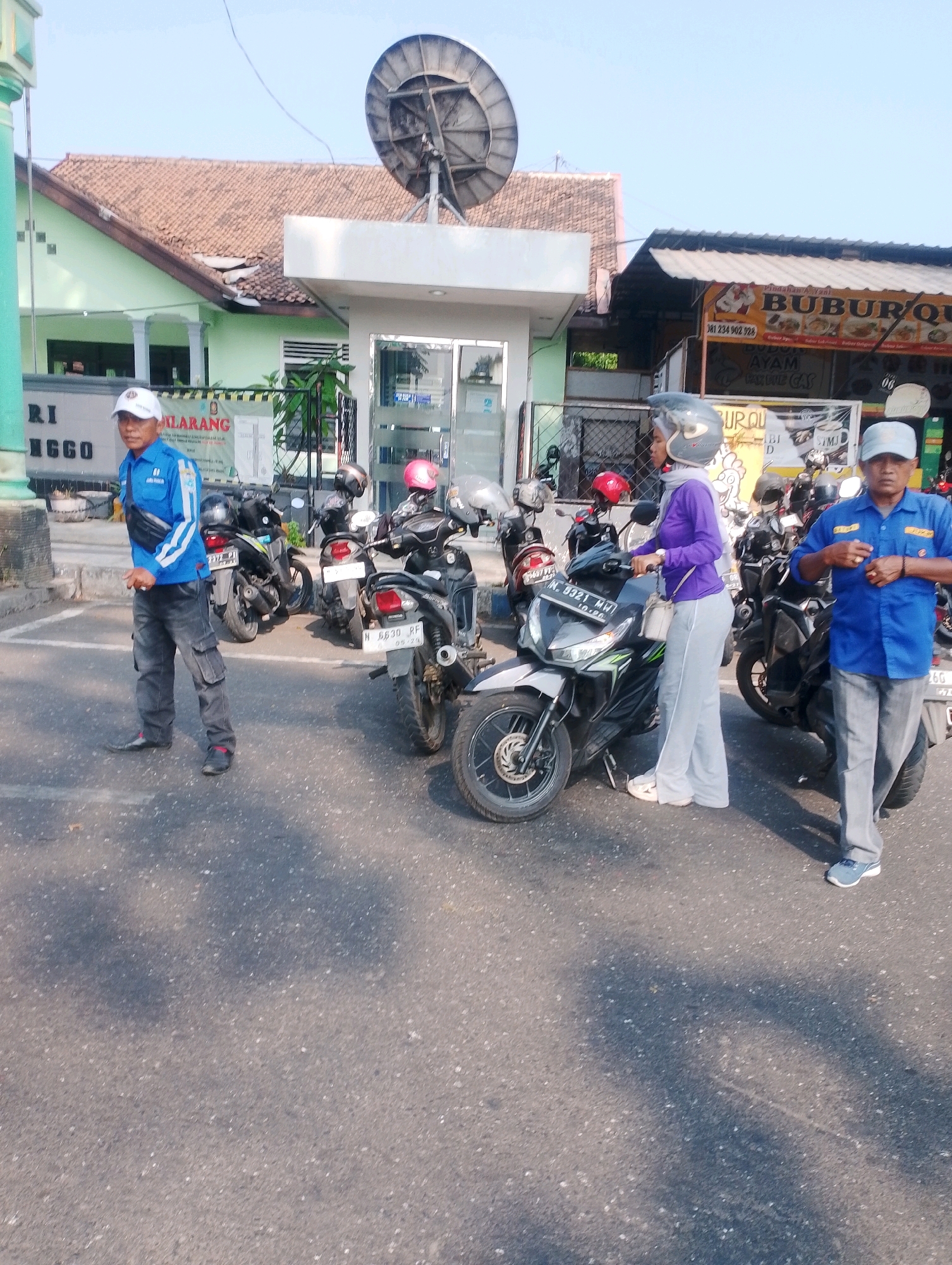 This image depicts an outdoor scene in the city of Probolinggo, Indonesia, likely near a commercial establishment. In the foreground, two men and one woman are present, each with distinct attire. The man on the left wears a blue long-sleeved shirt with white stripes on the sleeves, dark pants, and a white cap. He has a black waist bag slung across his chest. The woman in the middle is wearing a purple hijab and a purple long-sleeved shirt, paired with light gray pants and white shoes. She has a light-colored purse slung over her shoulder and is wearing a helmet. The man on the right is dressed in a bright blue short-sleeved shirt with yellow accents, gray pants, and blue and gray sneakers. He is also wearing a light-colored cap.
Several motorcycles are parked in the background, suggesting a parking area. One motorcycle in the center is black and silver, with a license plate starting with "L 6921 MW". Another motorcycle behind it is black, and to its right is a red motorcycle with a pink helmet on it. Further to the right, another black motorcycle is visible with a license plate starting with "N 6630 RF".
The environment includes a building with a light-colored facade and a dark-tiled roof in the background. A large satellite dish is mounted on a pole in front of this building. To the right of this structure, a smaller, open-air stall with a banner displaying text and images of food is visible, with the word "BUBUR" prominently featured. The ground is paved with asphalt. The sky above is clear and bright, indicating daytime. The overall impression is a casual street scene with people and parked vehicles.