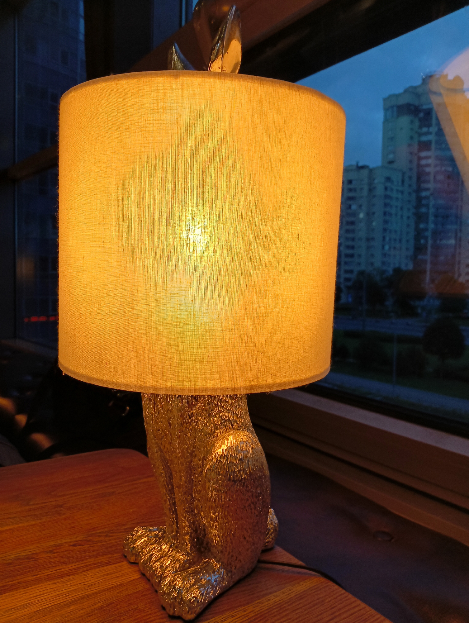 A table lamp with a textured gold base in the shape of a seated animal, possibly a cat, is positioned on a wooden table. The lamp has a cylindrical, fabric lampshade that emits a warm, yellow light. The lampshade is partially translucent, revealing the light bulb within. The lamp is situated near a large window, through which a city skyline is visible. The sky is overcast and a dusky blue, suggesting it is either early morning or evening. Buildings, some of which appear to be residential apartment blocks, are seen in the background, along with trees and what looks like a road. The image captures a scene indoors, with the lamp providing an intimate light source against the backdrop of the urban environment outside. The specific location in Saint Petersburg, Russia, cannot be determined from the visual information alone, as the image lacks any distinguishing landmarks or signage that would definitively identify the area.